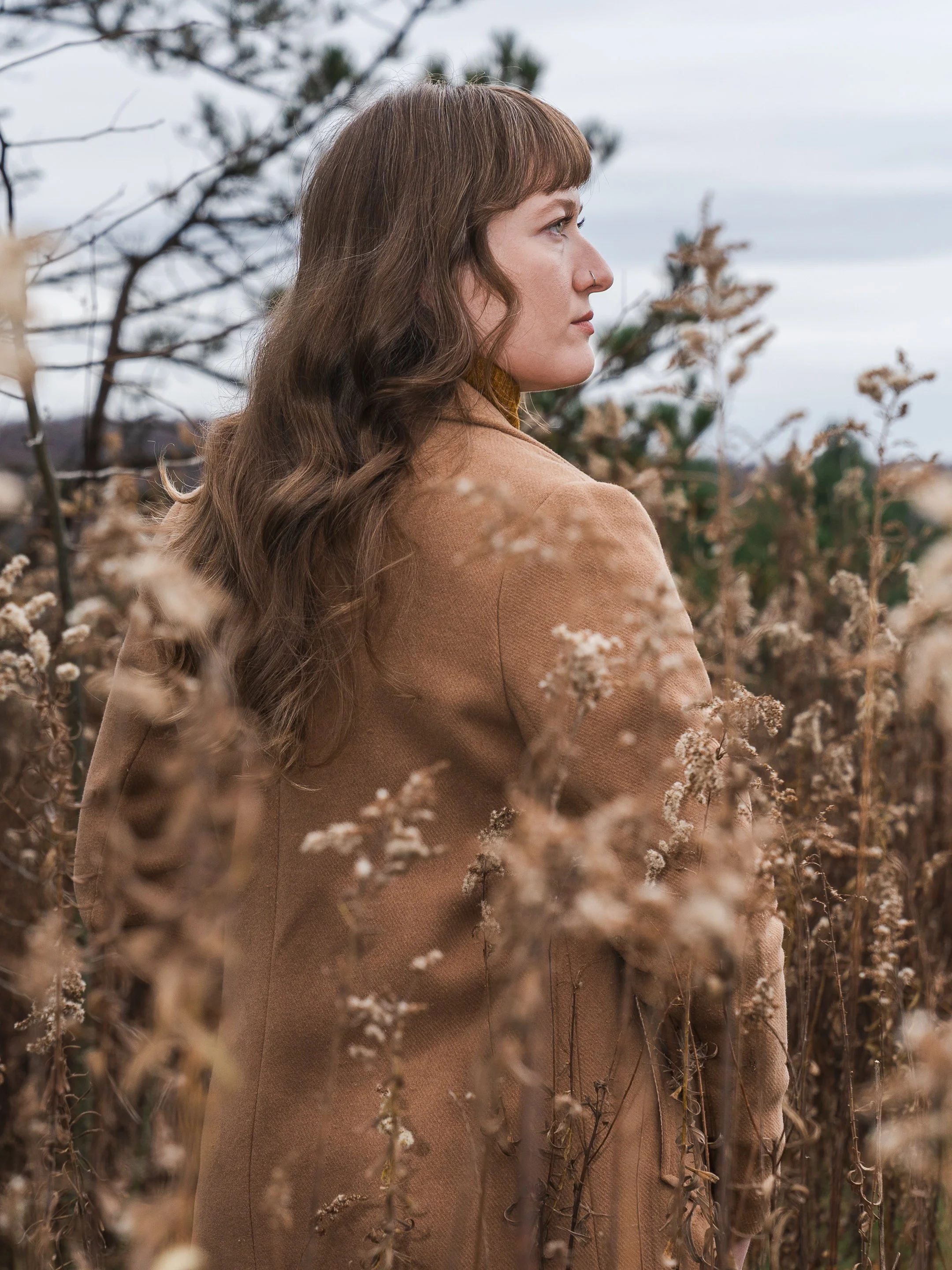A woman with long wavy brown hair and a nose ring looking to the right while standing among dry plants outdoors.