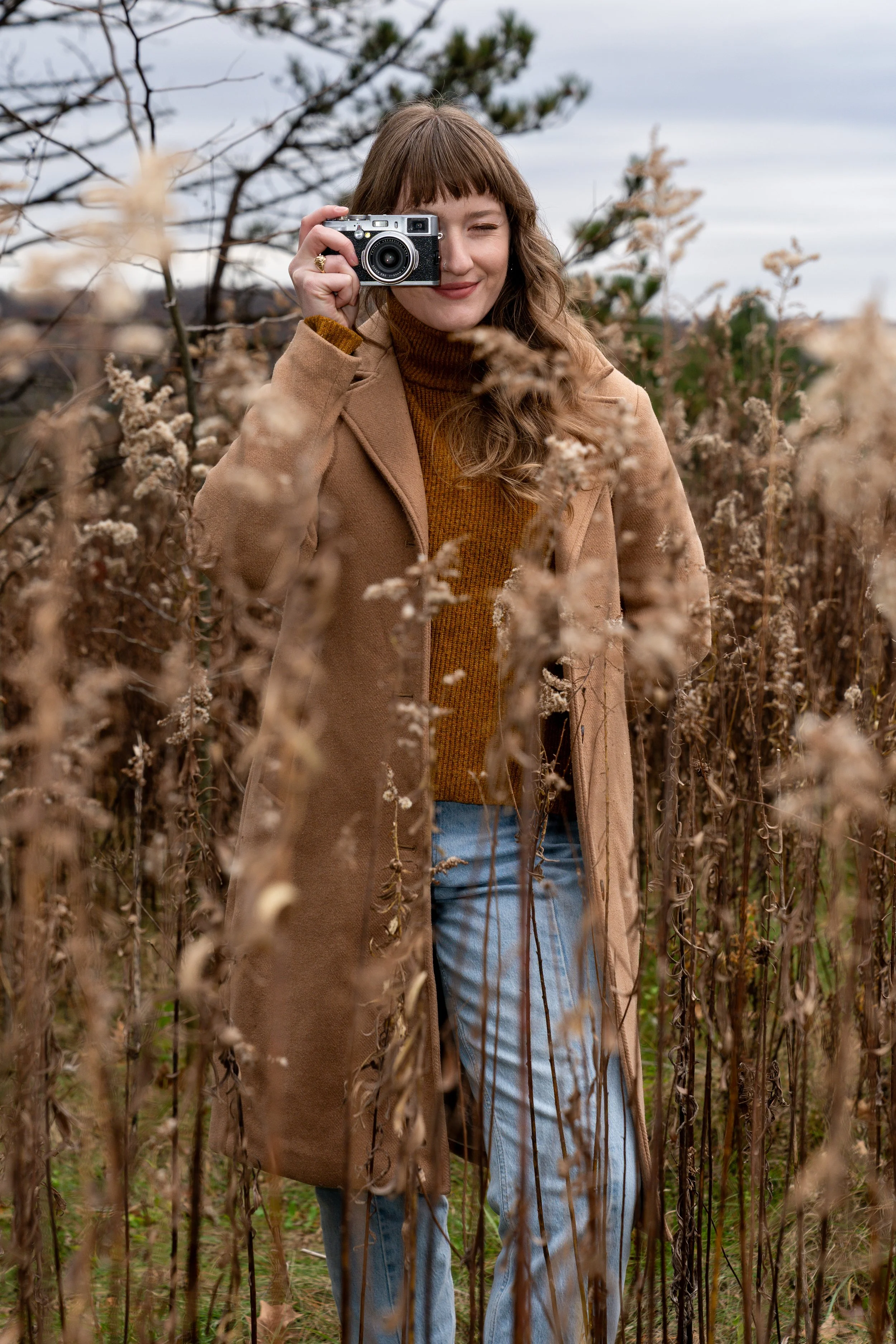 A woman with light brown hair and bangs, wearing a brown coat over a mustard-colored sweater, stands among dried plants outdoors on a cloudy day, holding a vintage camera up to her face and smiling.