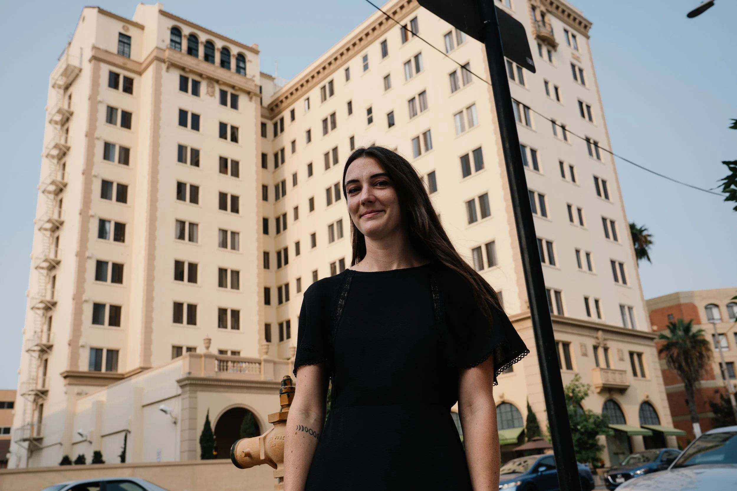 A woman with long dark hair and a black dress standing outdoors in front of a large cream-colored building with multiple windows, a fire hydrant and cars visible in the background.