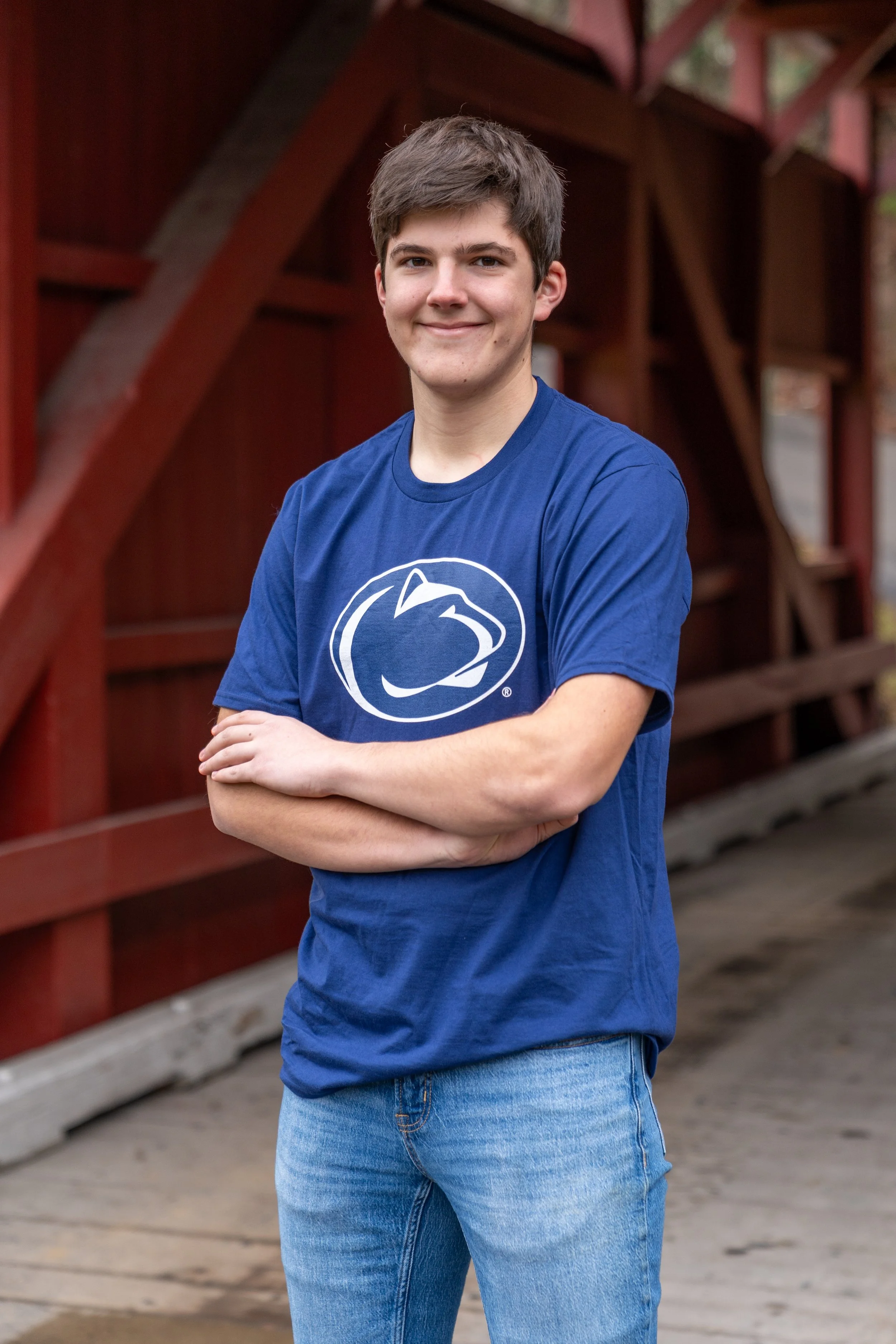 A young woman with short dark hair, smiling, wearing a blue Penn State Nittany Lions T-shirt, standing outdoors with arms crossed in front of a red wooden structure.