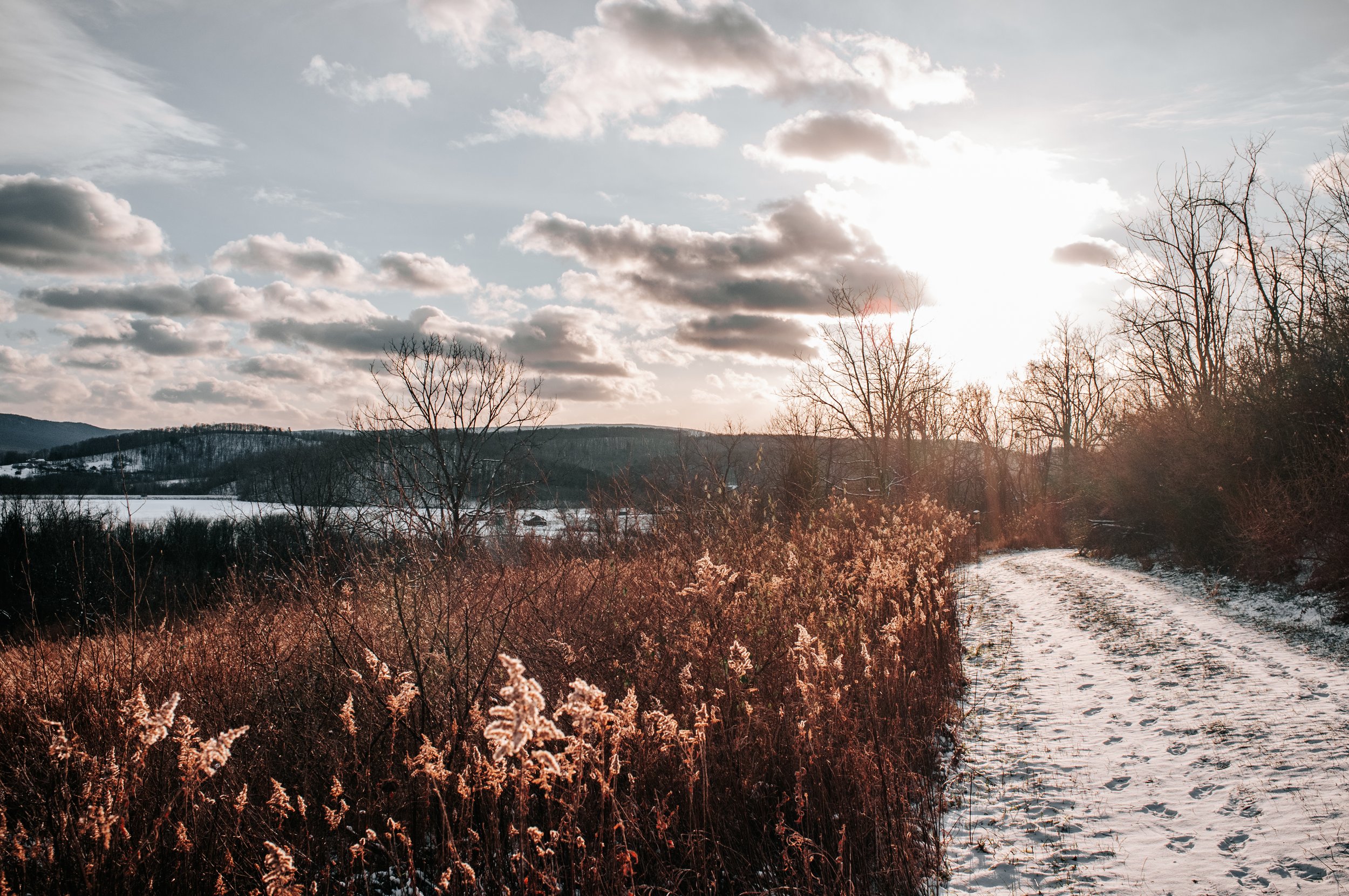 Sunset over a snowy rural landscape with leafless trees, shrubs, a dirt path, and distant hills.