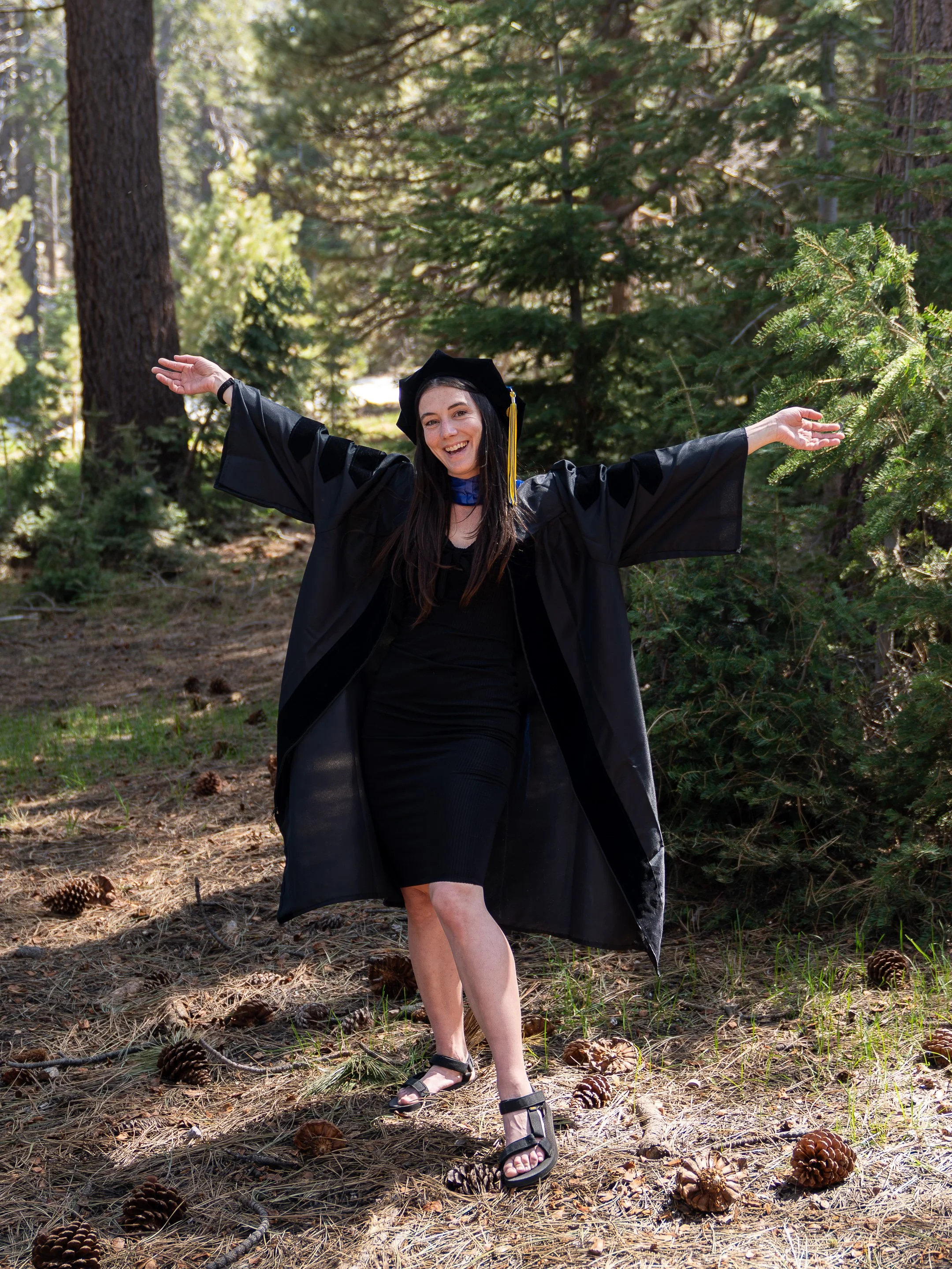 A woman in a graduation cap and gown poses joyfully with arms outstretched in a forest, surrounded by pinecones and pine needles.