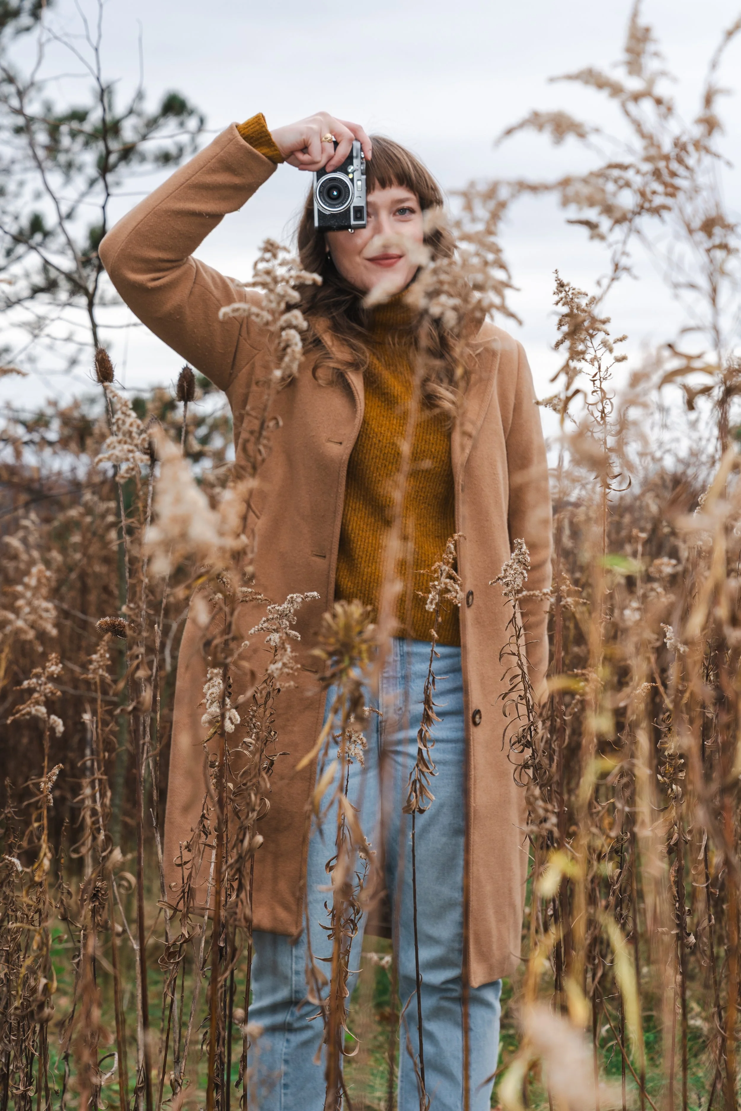 A woman with brown hair in a brown coat and blue jeans standing amidst tall dried plants, holding a camera up to her face for a selfie or photo.