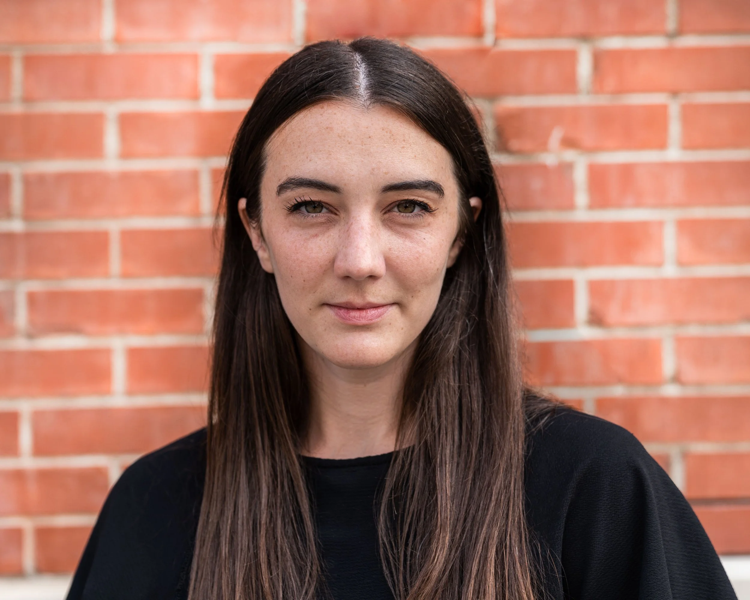 Close-up of a young woman with long brown hair, freckles, and hazel eyes standing in front of a red brick wall