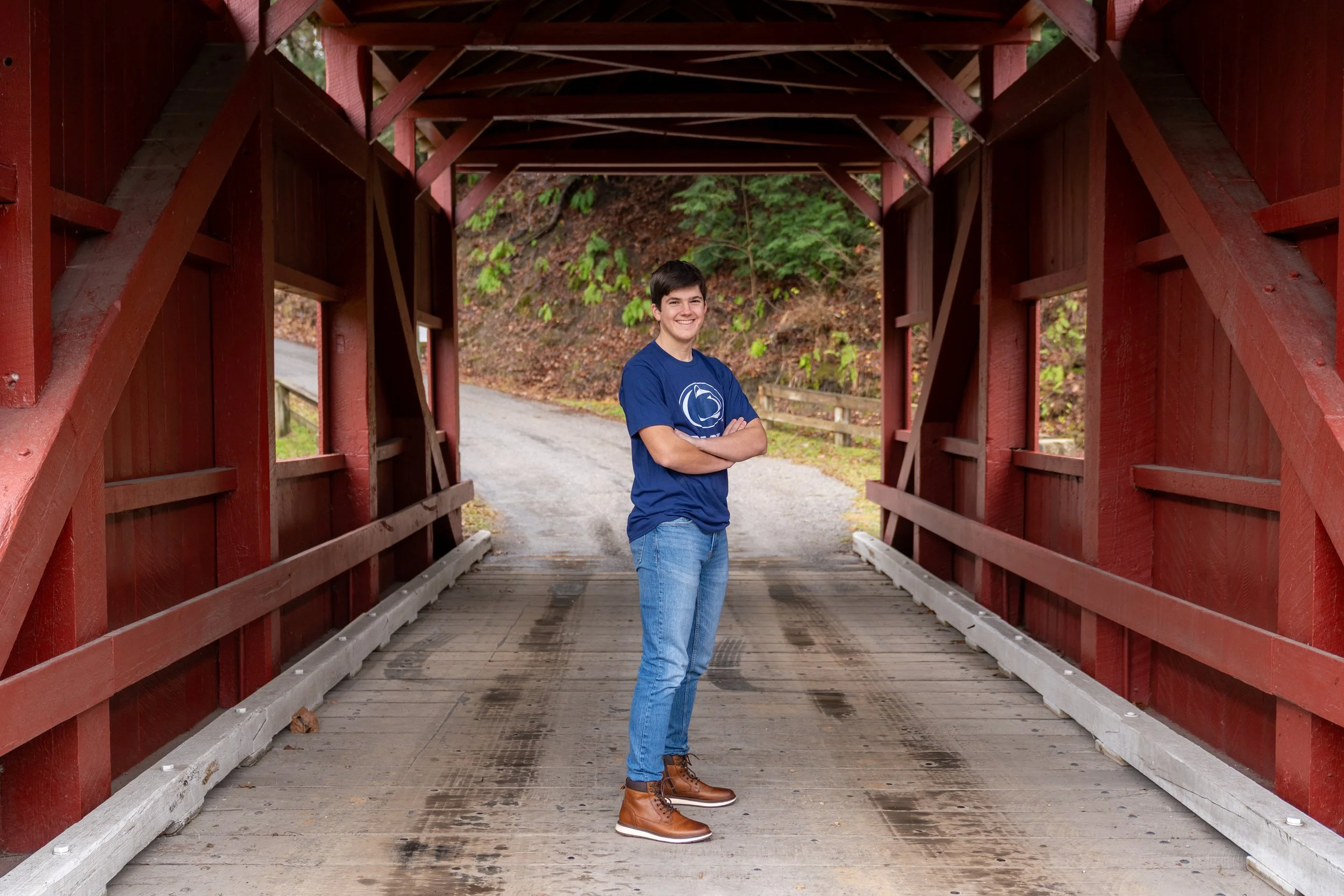 Young man standing confidently inside a red wooden covered bridge, smiling with arms crossed, on a rainy day.