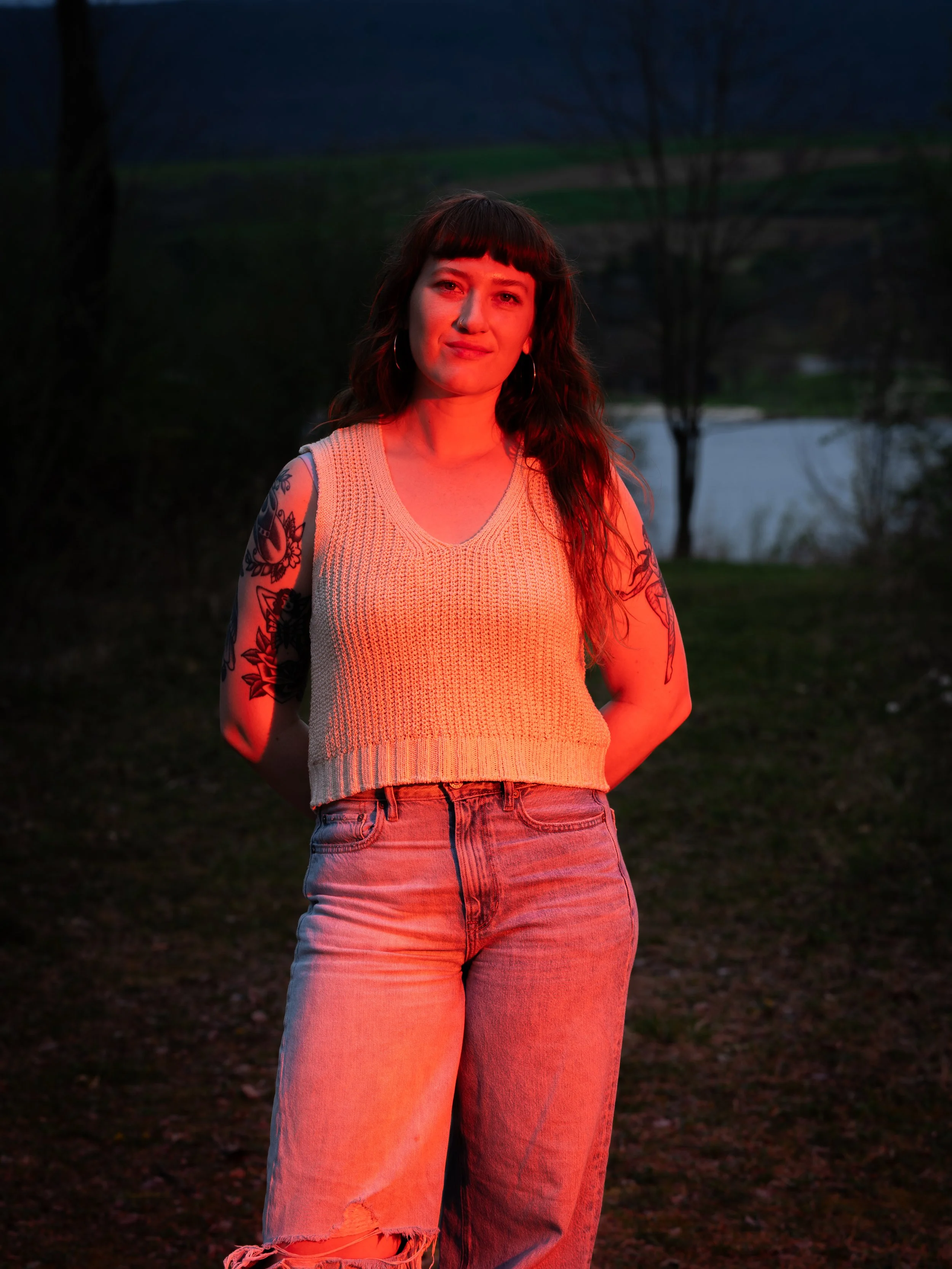 A woman standing outdoors at dusk or night, illuminated by a red light, with trees and a body of water in the background. She has tattoos on her arms, wears a sleeveless beige knit top, earrings, and light-wash ripped jeans.