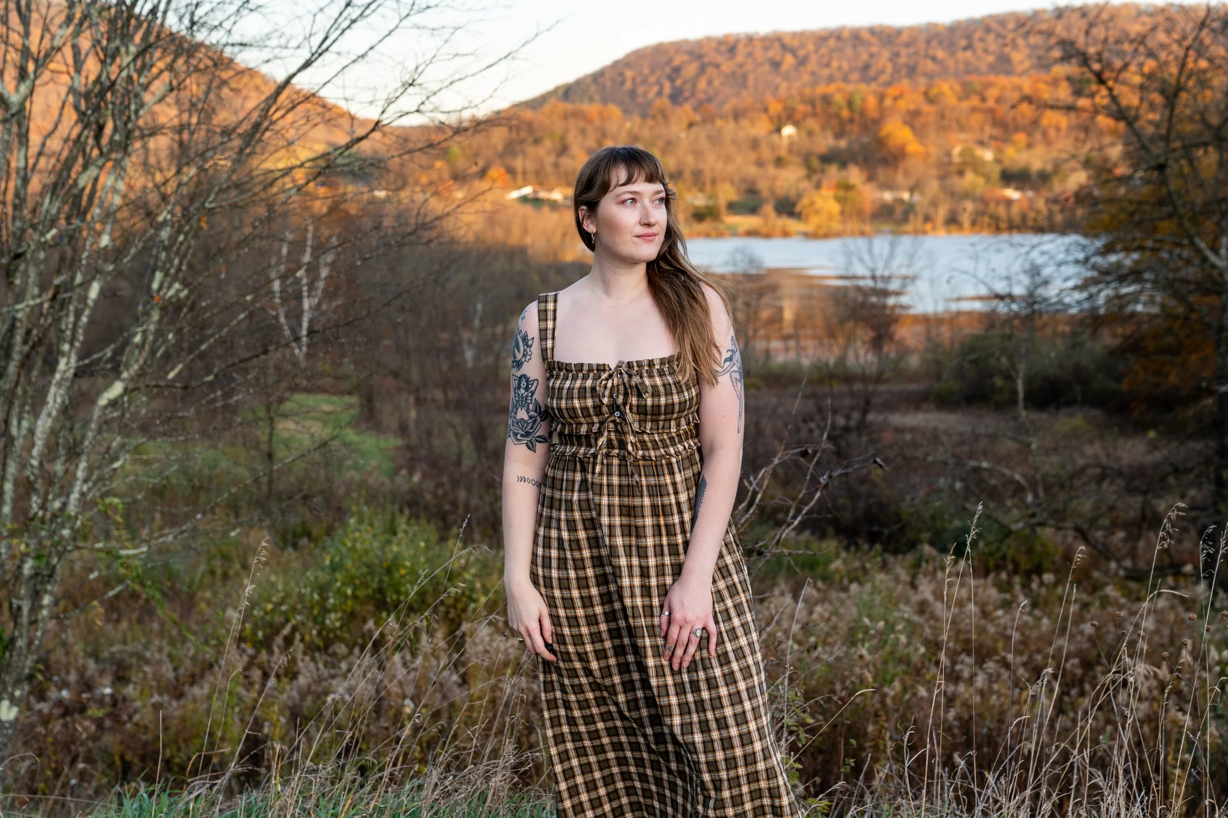 A young woman with tattoos on her arms wearing a plaid dress standing outdoors among trees and bushes, with a lake and rolling hills with autumn foliage in the background during sunset.