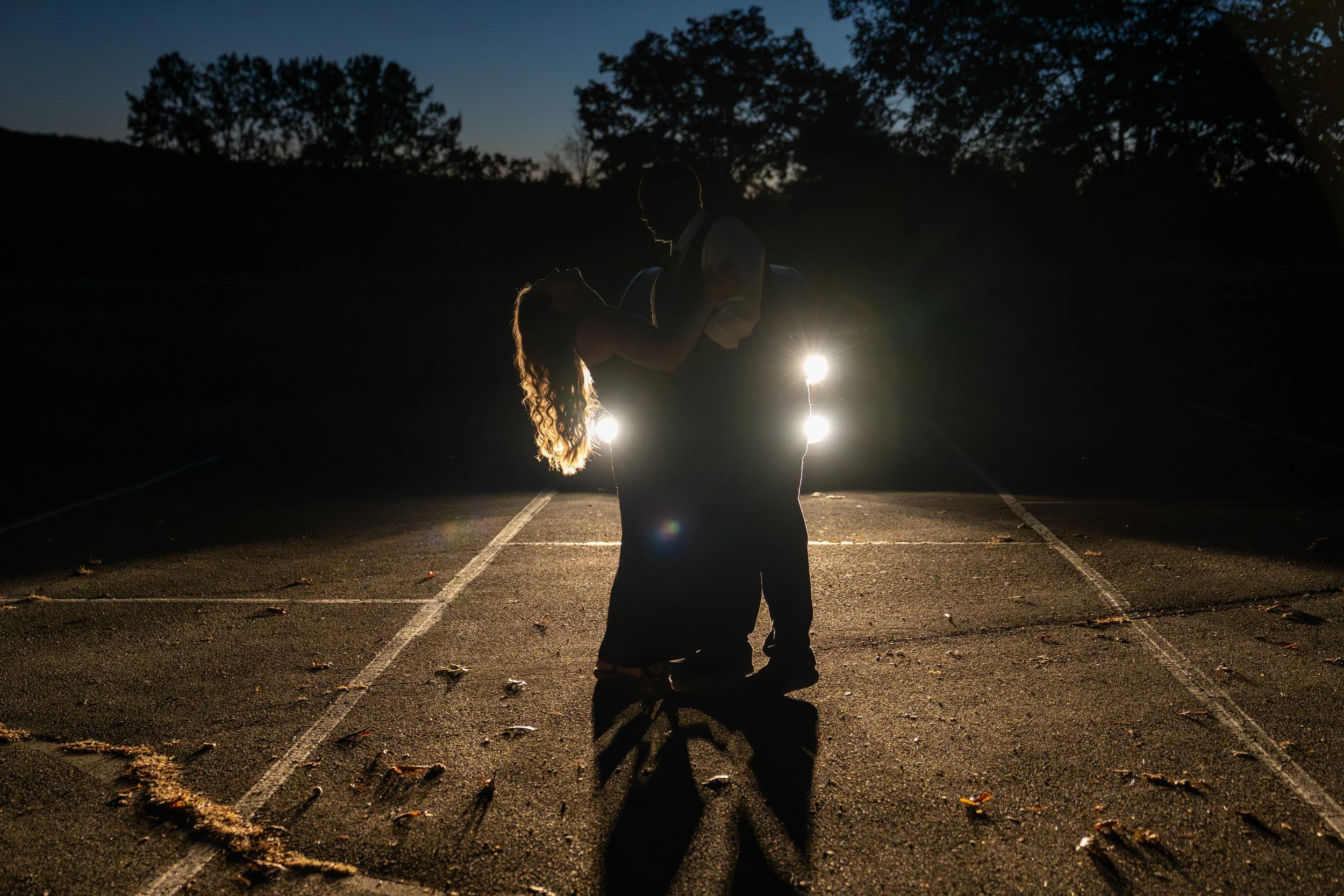 A silhouette of a couple dancing in an empty parking lot at night, with trees in the background and the woman’s long hair illuminated by lights.