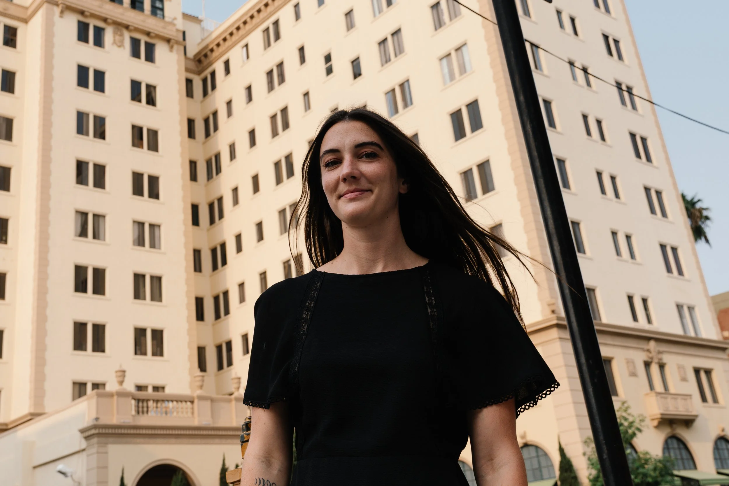 A woman with dark hair smiling slightly, wearing a black top, standing outdoors in front of a tall building with multiple windows, and a black pole nearby.