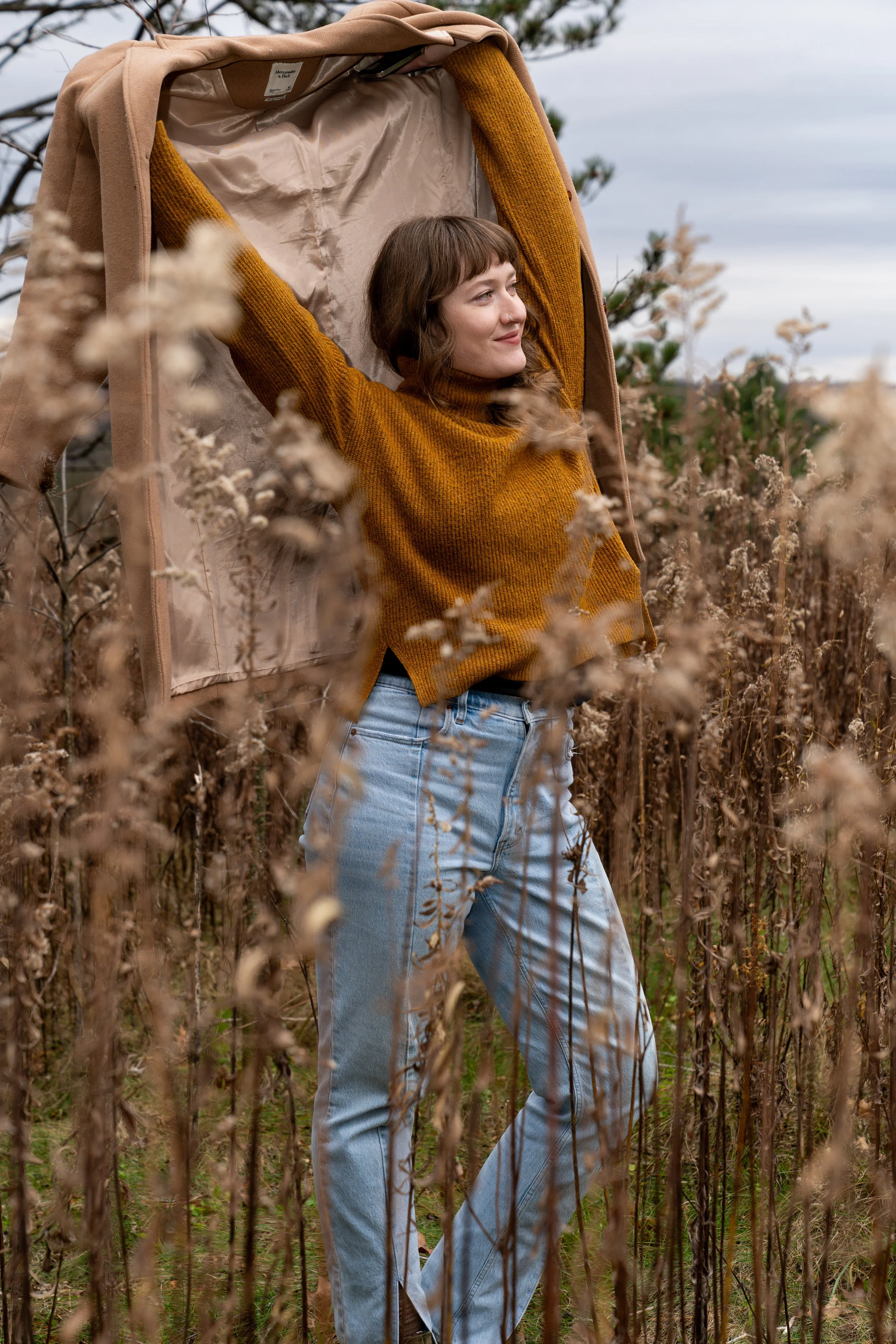 A young woman with brown hair in a bob haircut, wearing a rust-colored sweater and light blue jeans, standing in a field of dried plants or weeds outdoors on a cloudy day with her arms raised and resting on the inside of a beige canopy or tent.