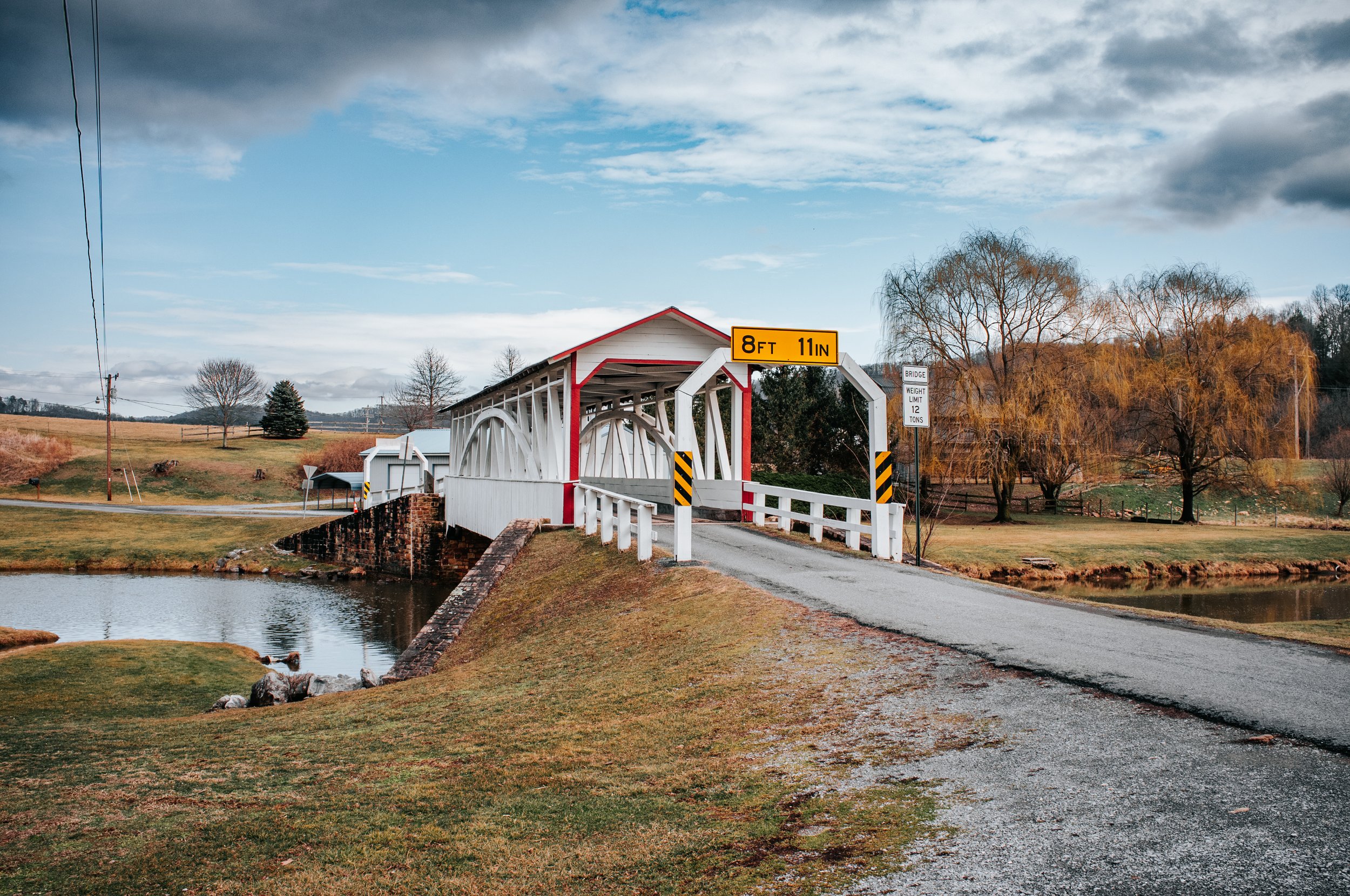 A small white covered bridge over a pond, with a gravel road leading onto it. Sign indicating 8 feet 11 inches height limit. Trees and grassy fields in the background under partly cloudy sky.