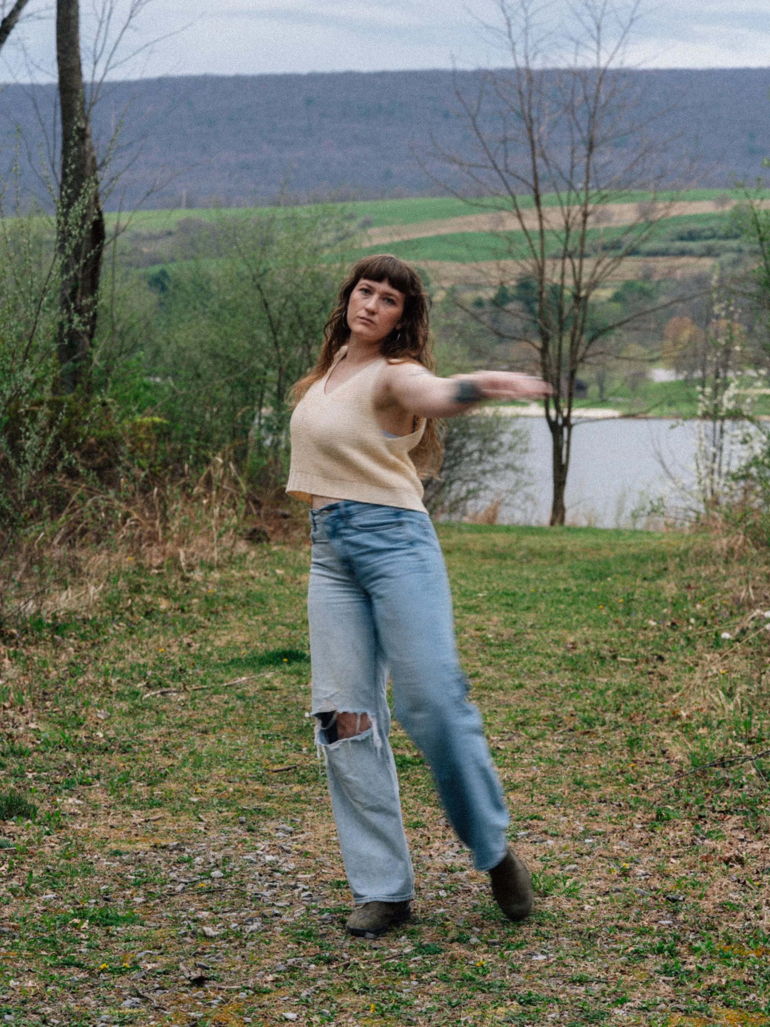 A woman standing on a grassy trail near a lake, with hills and leafless trees in the background, extending her right arm outward.