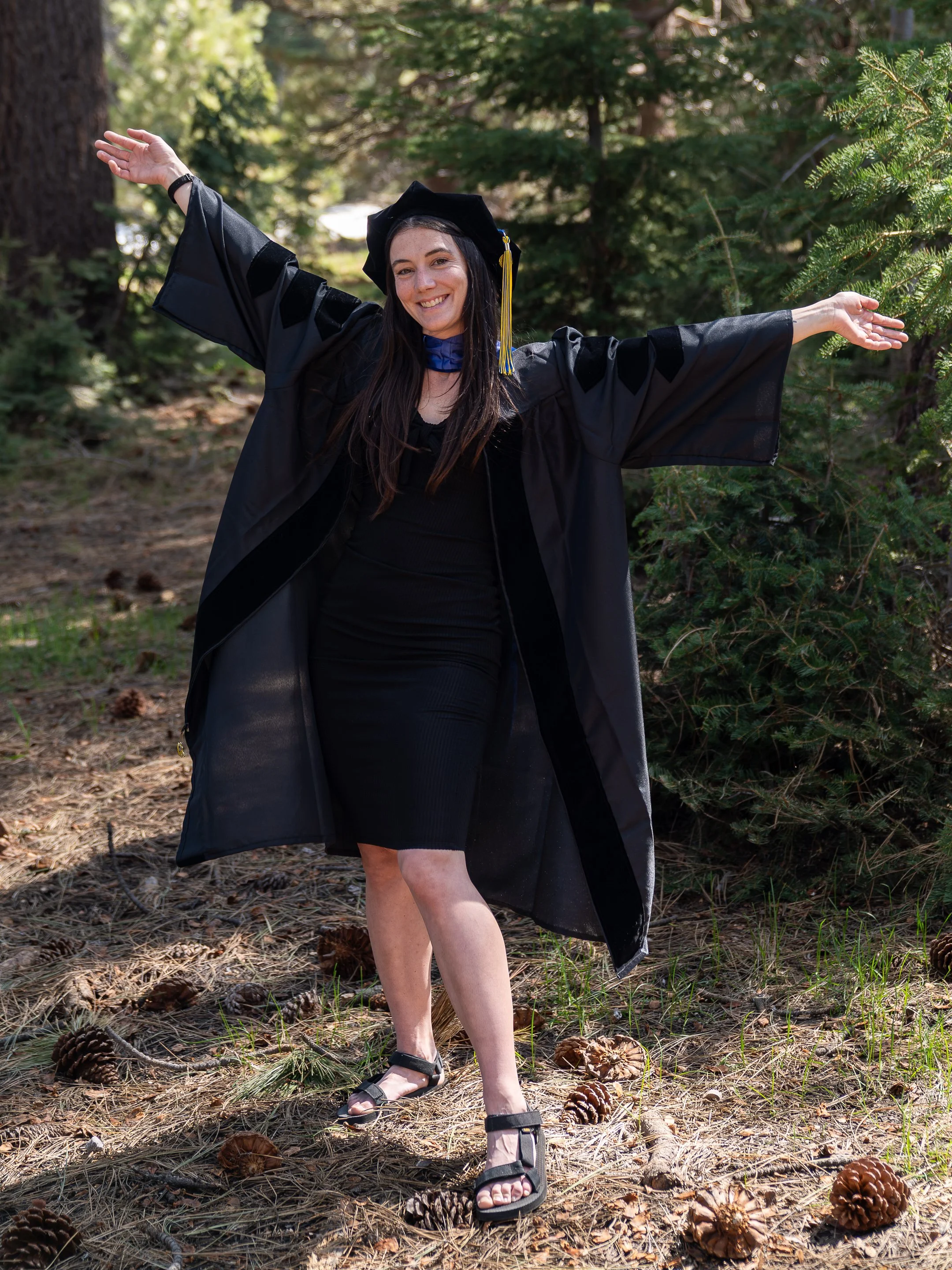 A woman in a black graduation gown and cap standing outdoors in a forest, smiling with arms outstretched, surrounded by pinecones and pine needles.