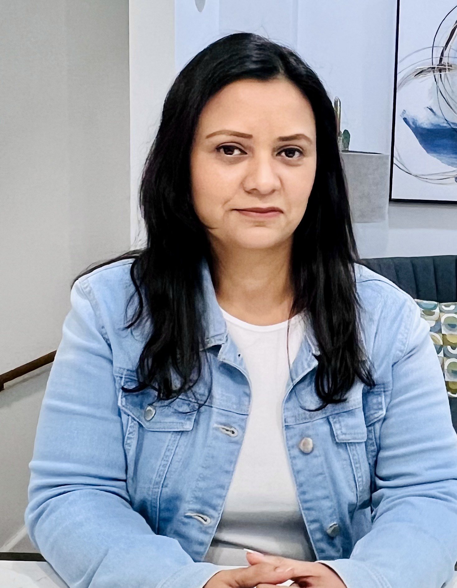 A woman with long black hair sitting at a table in a modern office or cafe, with a abstract art piece on the wall behind her.
