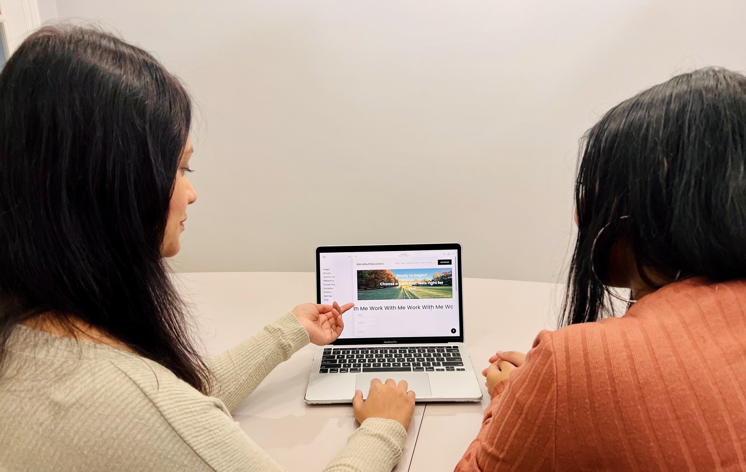 Two women sit at a table looking at a computer screen with a website displayed. One woman is pointing at the screen, and the other is sitting with her hands clasped. The background is a plain light-colored wall.