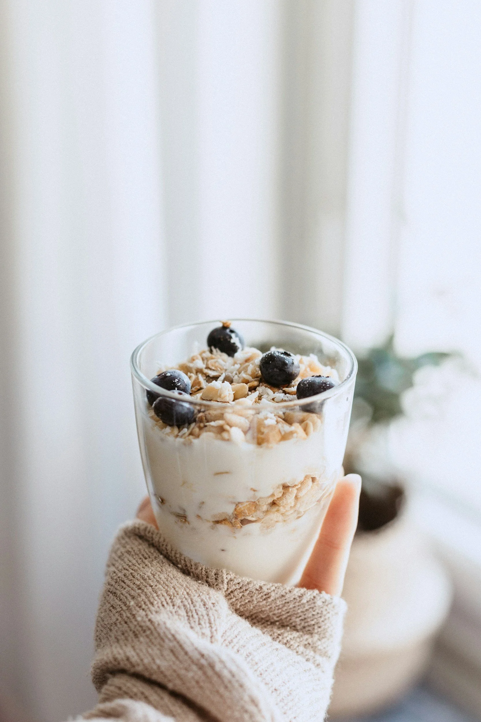 A hand wearing a beige knit glove holding a glass of layered yogurt with granola and blueberries near a blurred background of a white curtain and a potted plant.