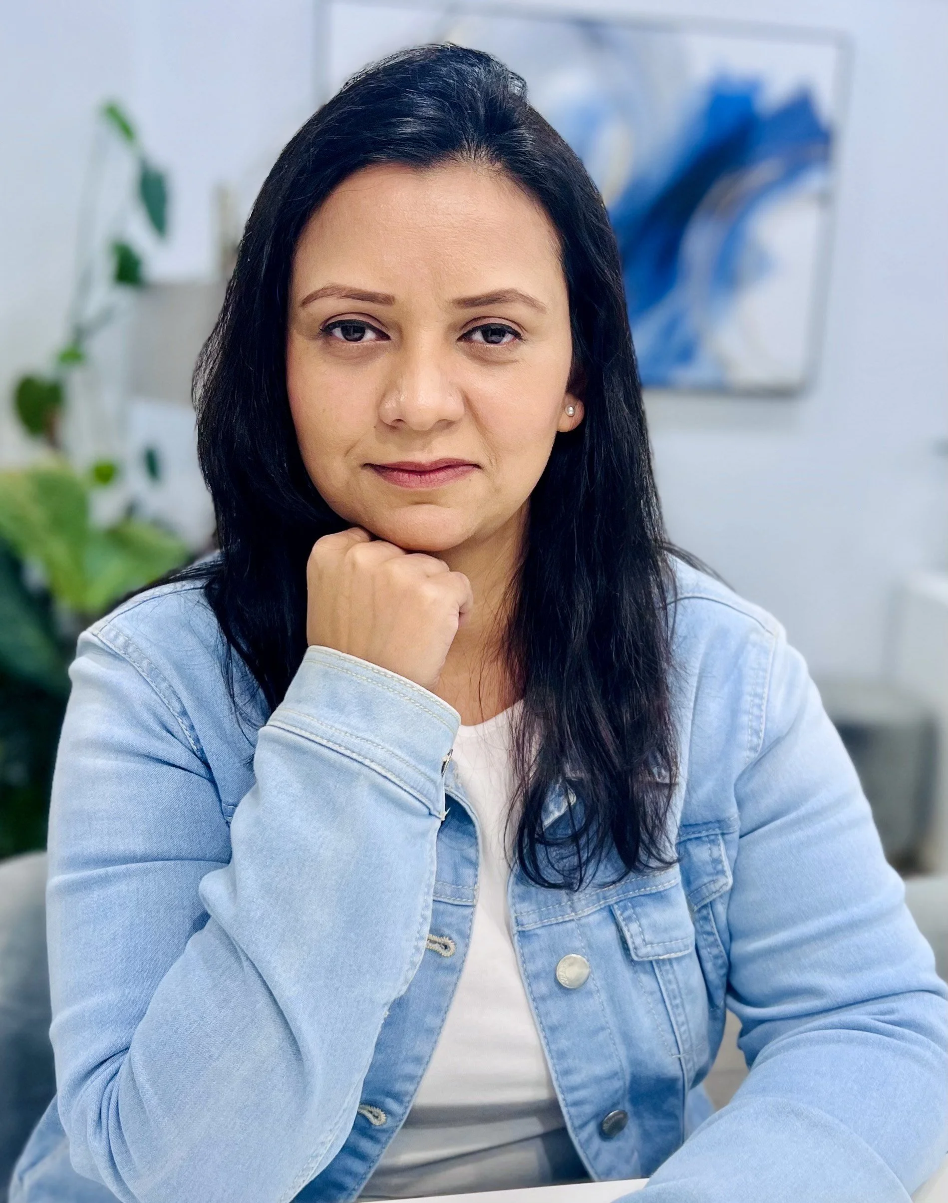 A woman with long black hair, resting her chin on her hand, looking confidently at the camera. She is wearing a light blue denim jacket in a modern, well-decorated indoor setting.