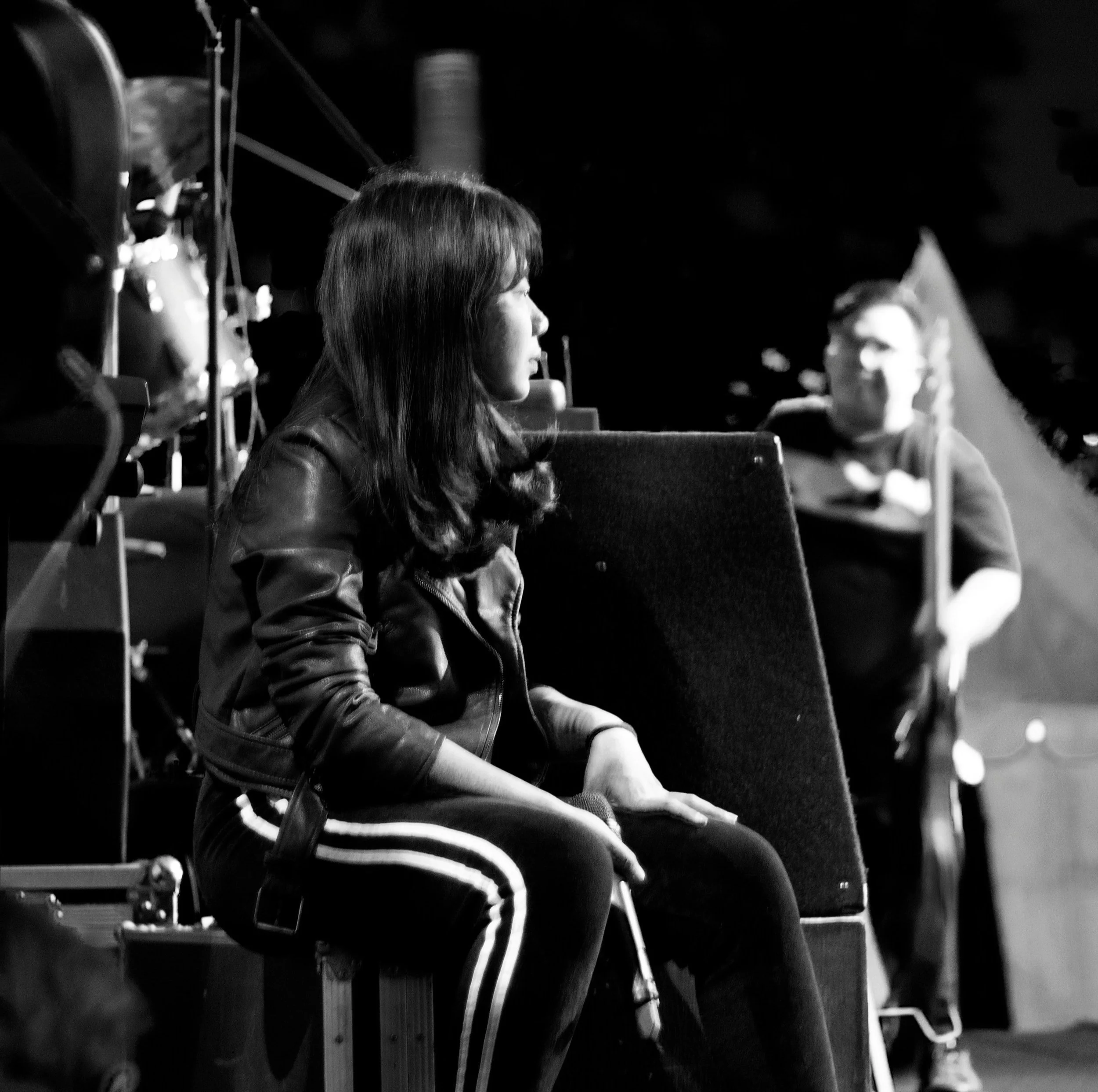A woman with long hair sitting on a bench, in profile view, wearing a leather jacket and striped pants, looking at a person in the background with a blurred face, possibly on a stage.