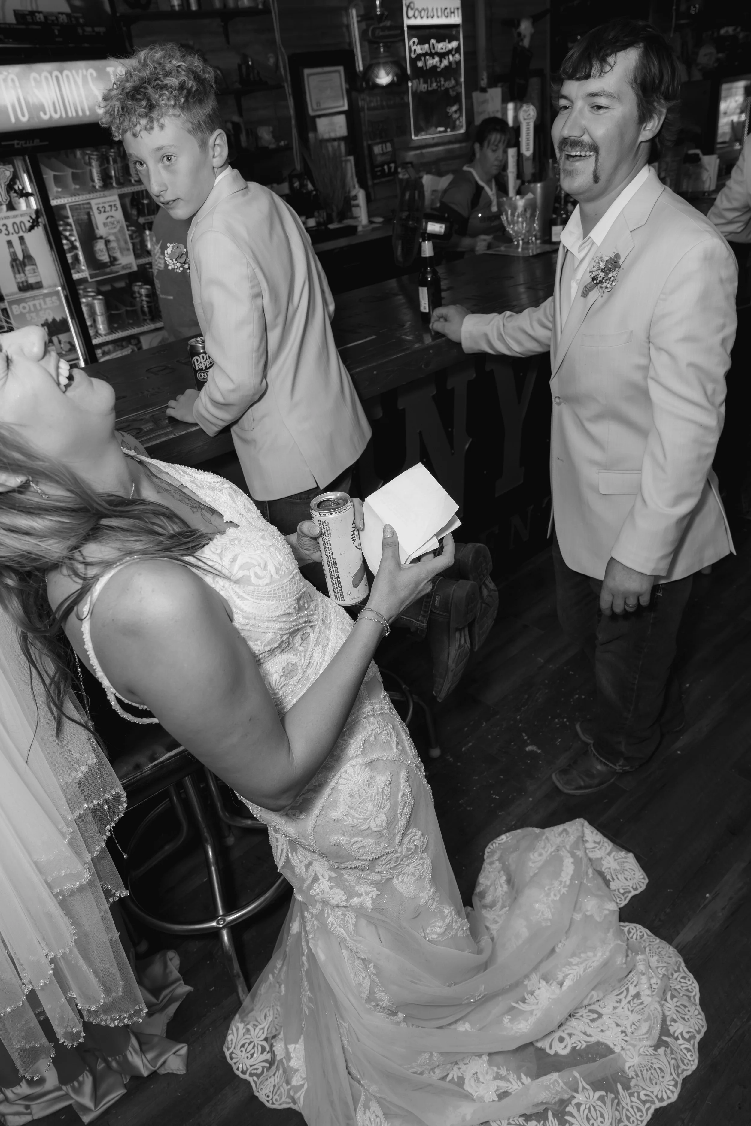 A woman in a wedding dress holding a can and a box, standing in a bar. A man in a light-colored suit with a boutonniere smiling at her. There is a young boy in a light blazer. The bar has a dark wooden counter, and a bartender and other people are visible in the background.
