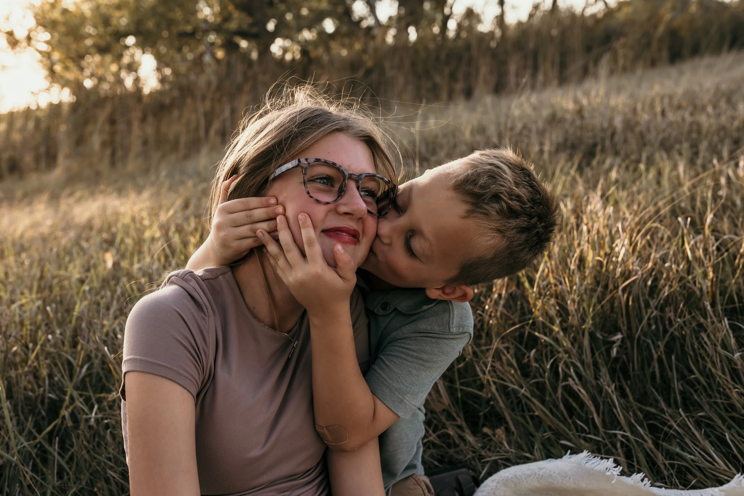 A young boy kisses his sister on the cheek, holding her face gently, as they sit in a field of tall grass during sunset. The girl wears glasses and a neutral-colored shirt, smiling softly.