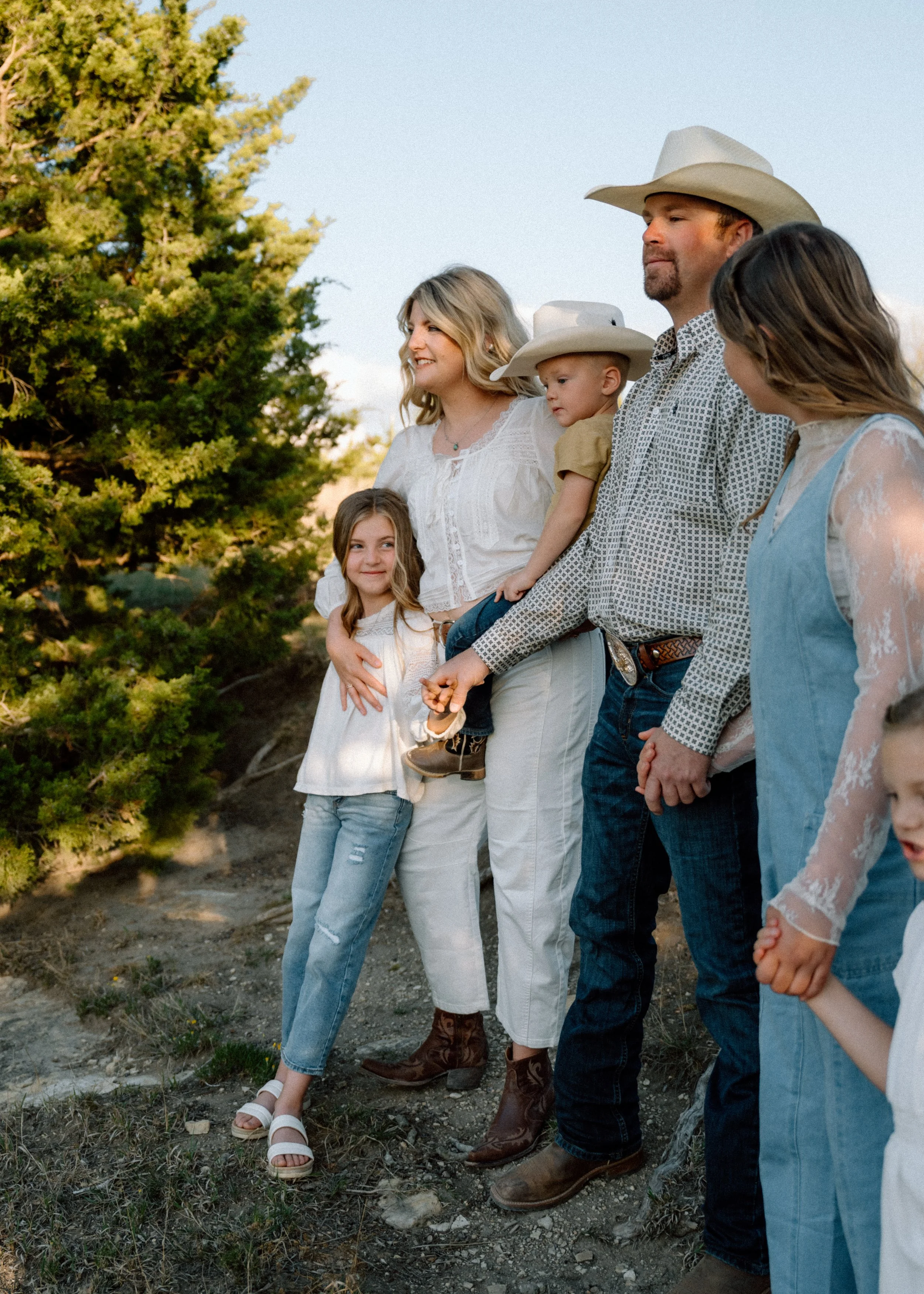 Family of six standing outdoors near trees, holding hands and smiling. They are dressed in casual, country-style clothing, with some wearing cowboy hats.