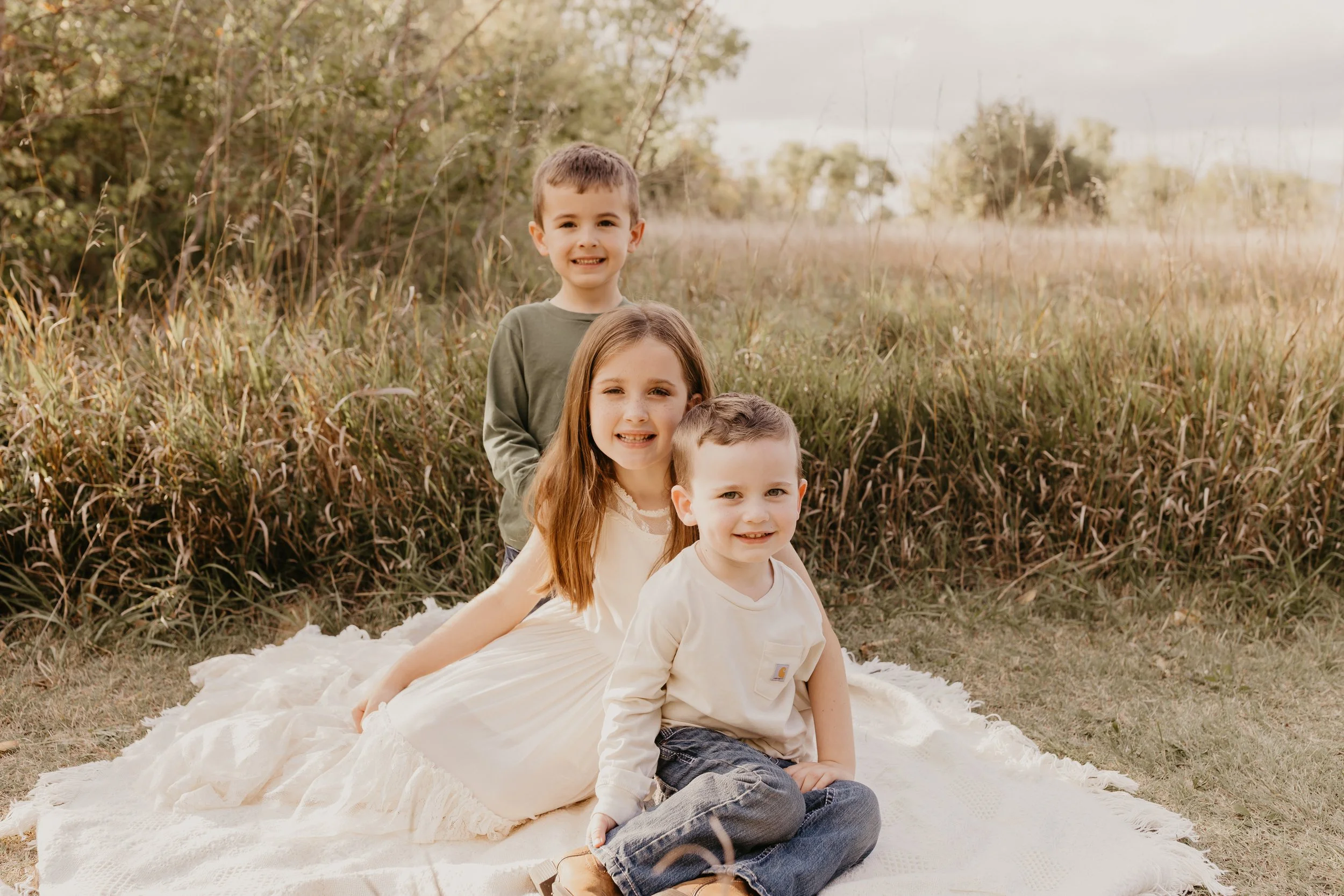 Three children outdoors, sitting on a blanket in a grassy field with trees and cloudy sky in the background.