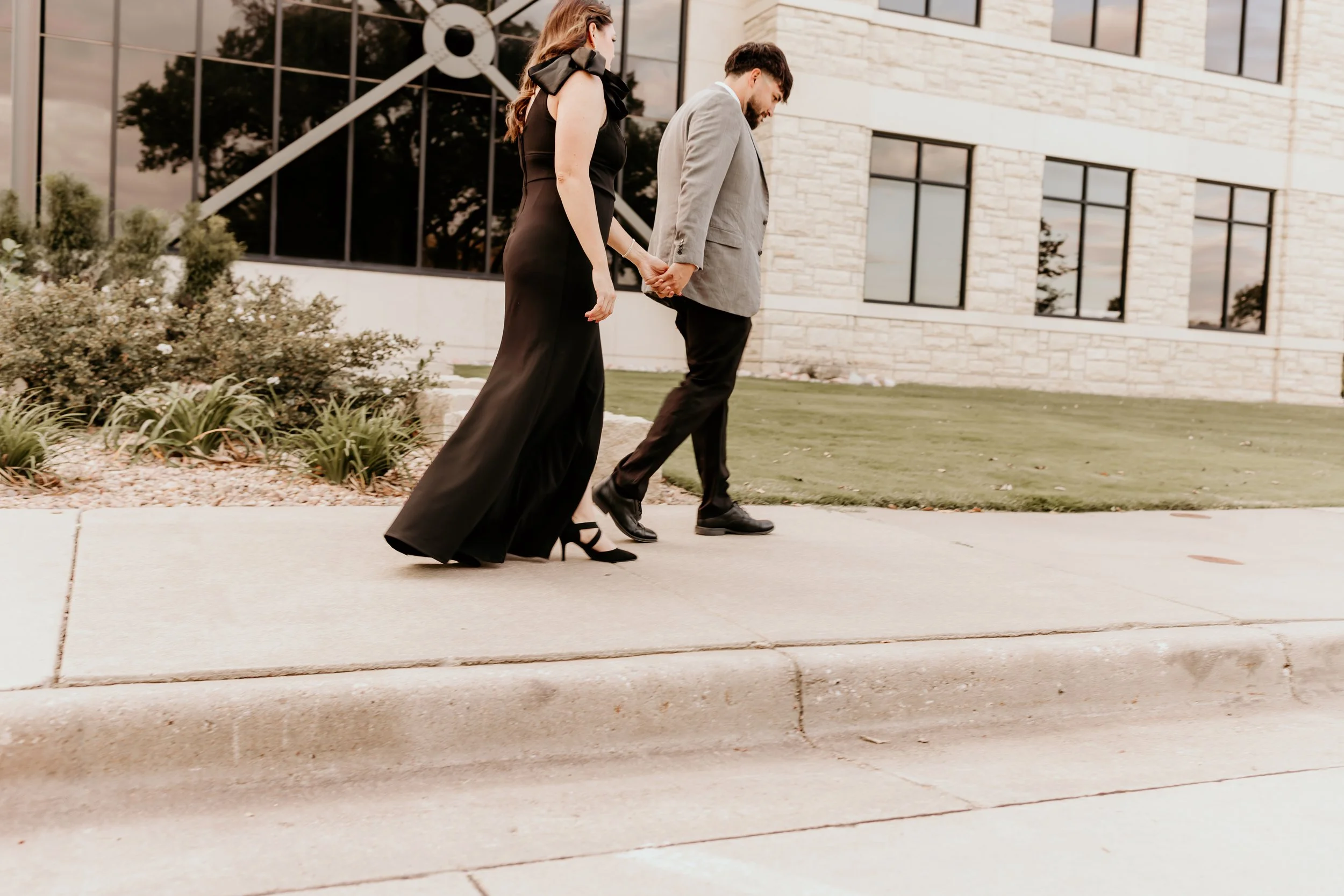 A couple holding hands and walking on a sidewalk outside a modern building during sunset.