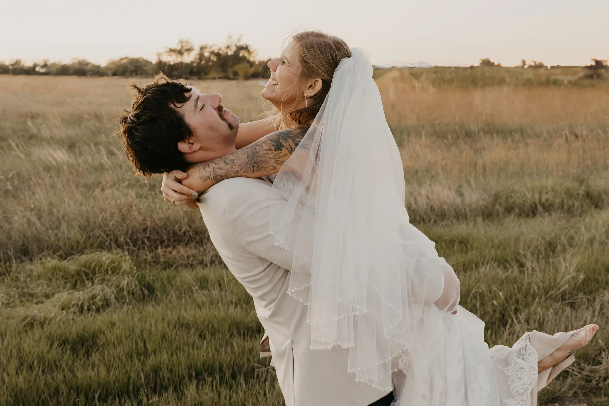 A groom in a white suit lifts a woman in a wedding dress with a veil, both smiling and looking at each other, in a grassy field during sunset.