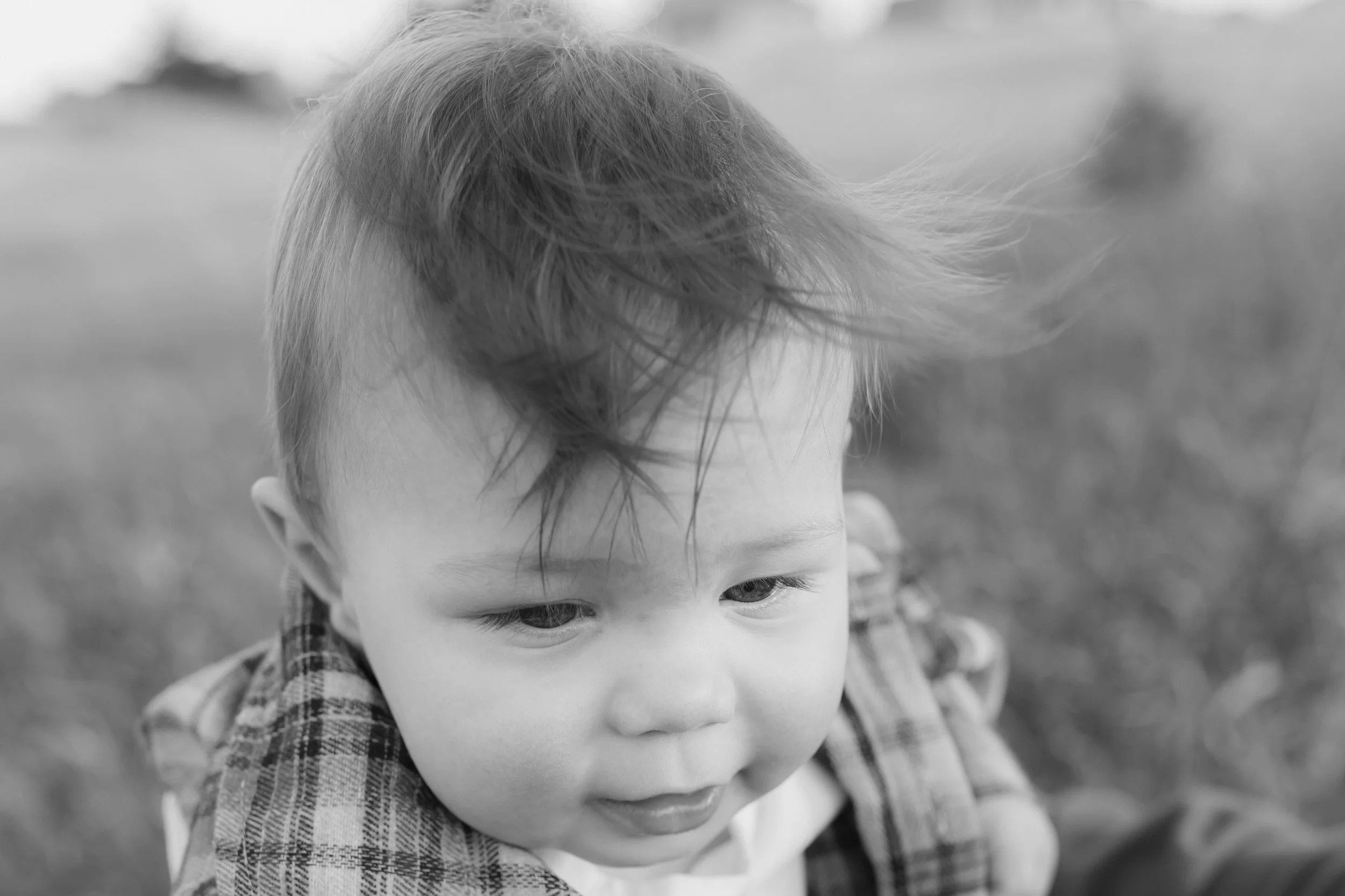 Close-up of a young child with tousled hair outdoors, wearing a plaid shirt, looking down thoughtfully.