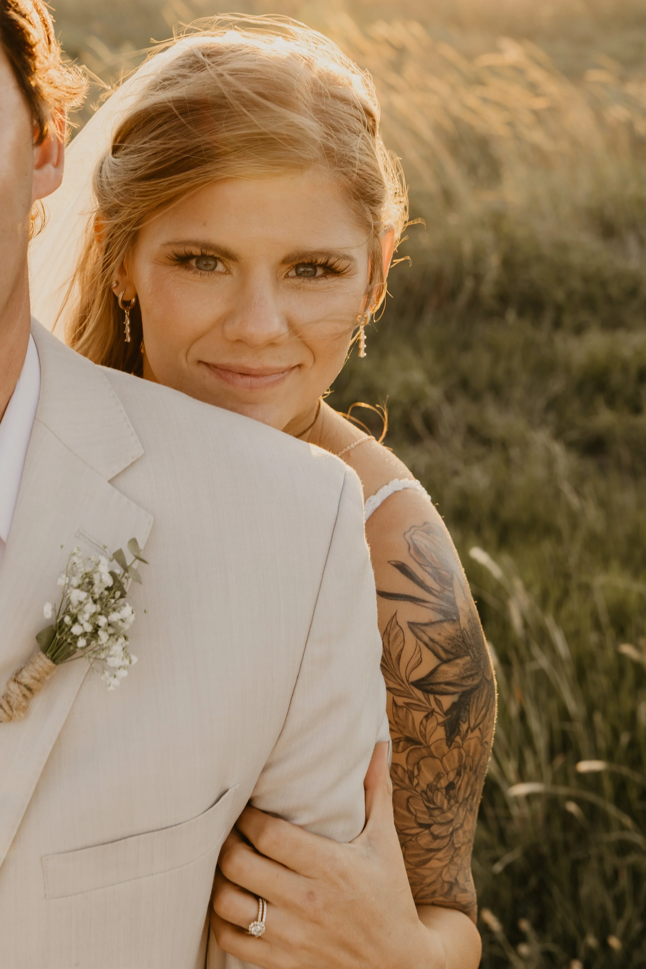 Close-up of a smiling bride with blonde hair, wearing earrings and a wedding dress with a tattooed arm, embracing her husband in a light-colored suit, outdoors during sunset.