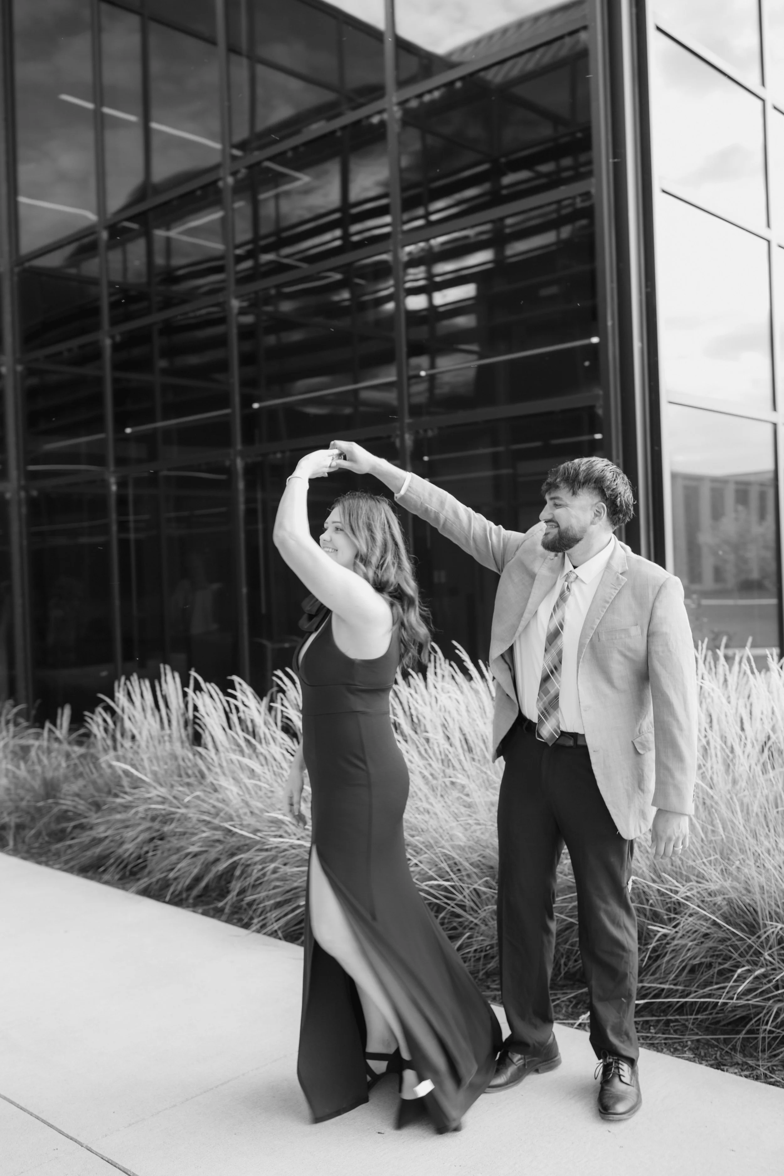A man and woman dancing together outdoors near a modern glass building with flowers in the background for an engagement session