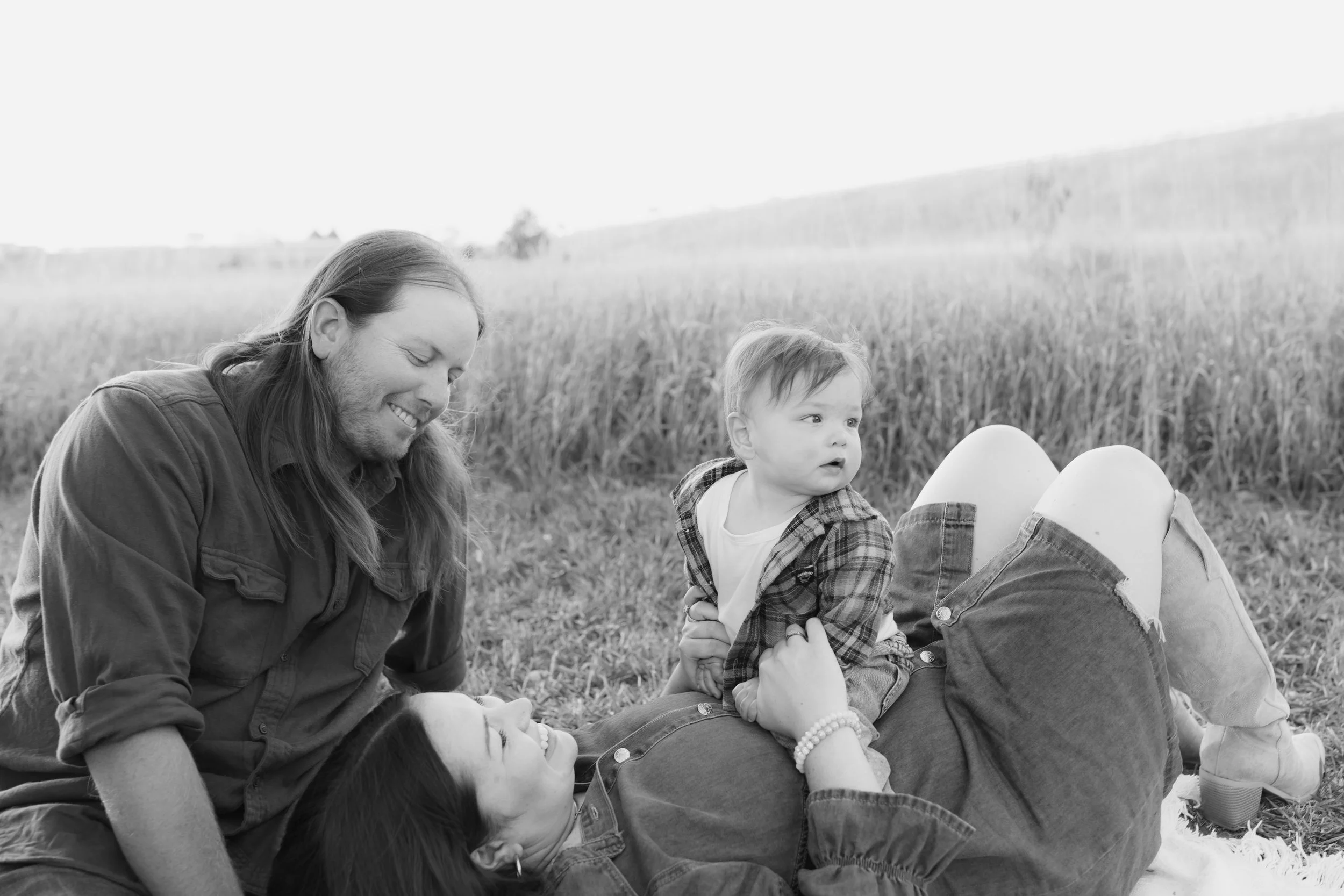 A happy family lying on the grass in a field, with a man, a woman, and a young child, smiling and playing together.
