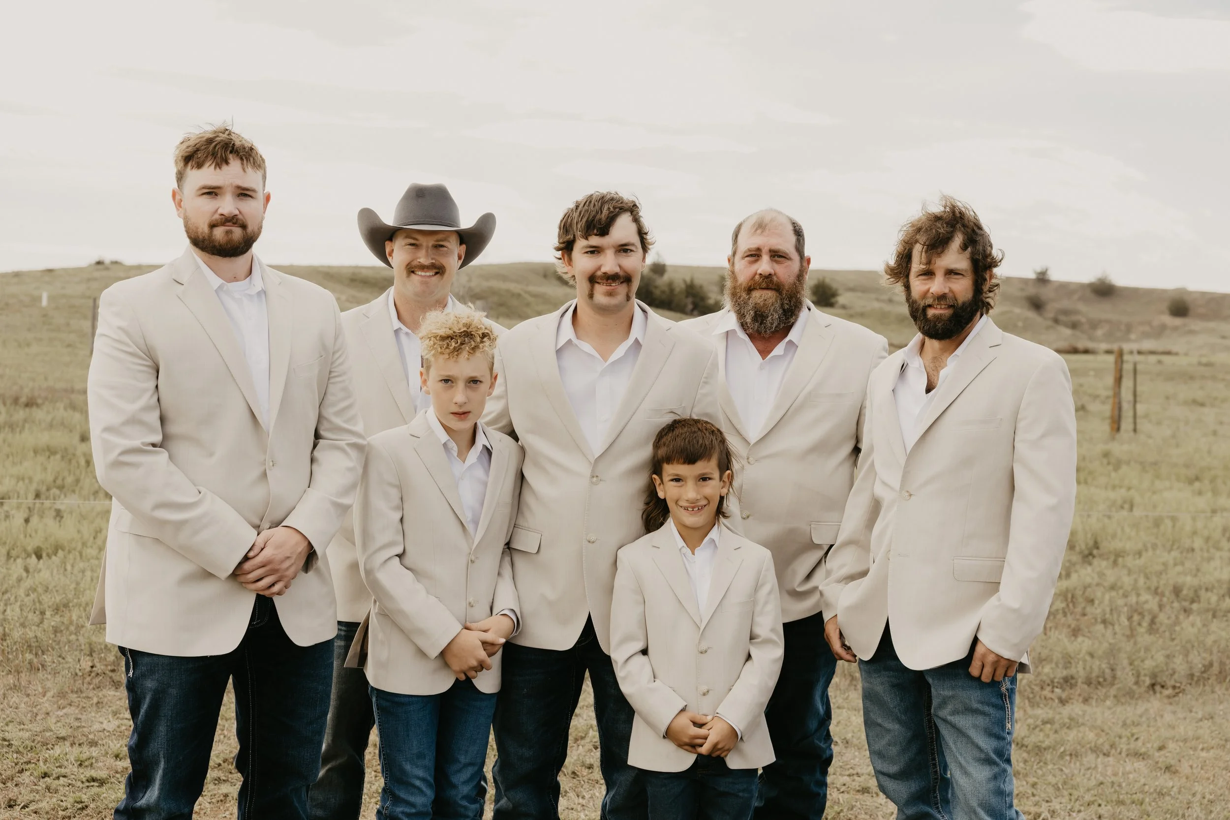 Group of five groomsmen and two boys standing outdoors in a field, all dressed in light-colored blazers and white shirts, with rolling hills and cloudy sky in the background.