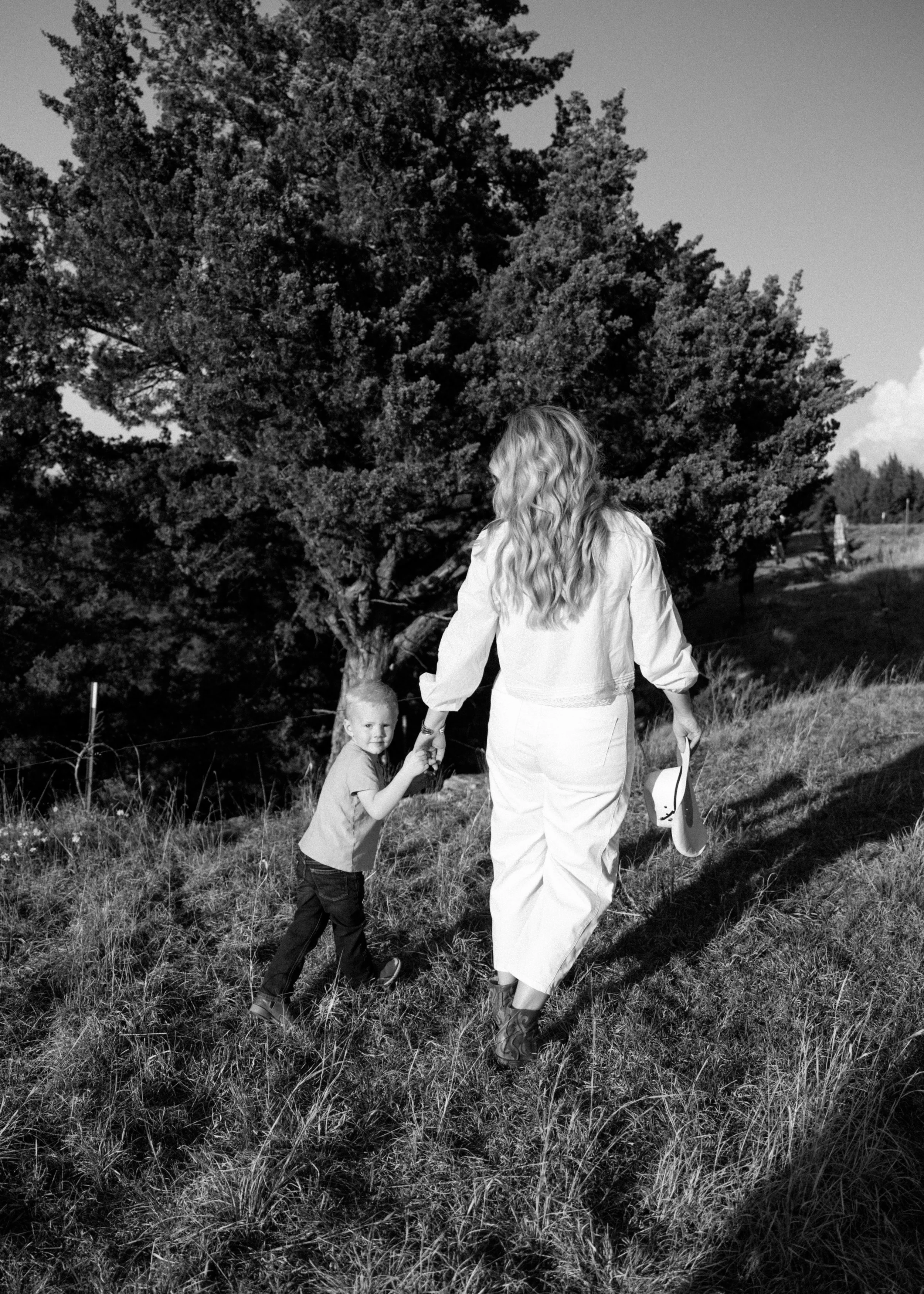 A woman with long, wavy hair walking on a grassy hillside holding a child's hand, with trees in the background.