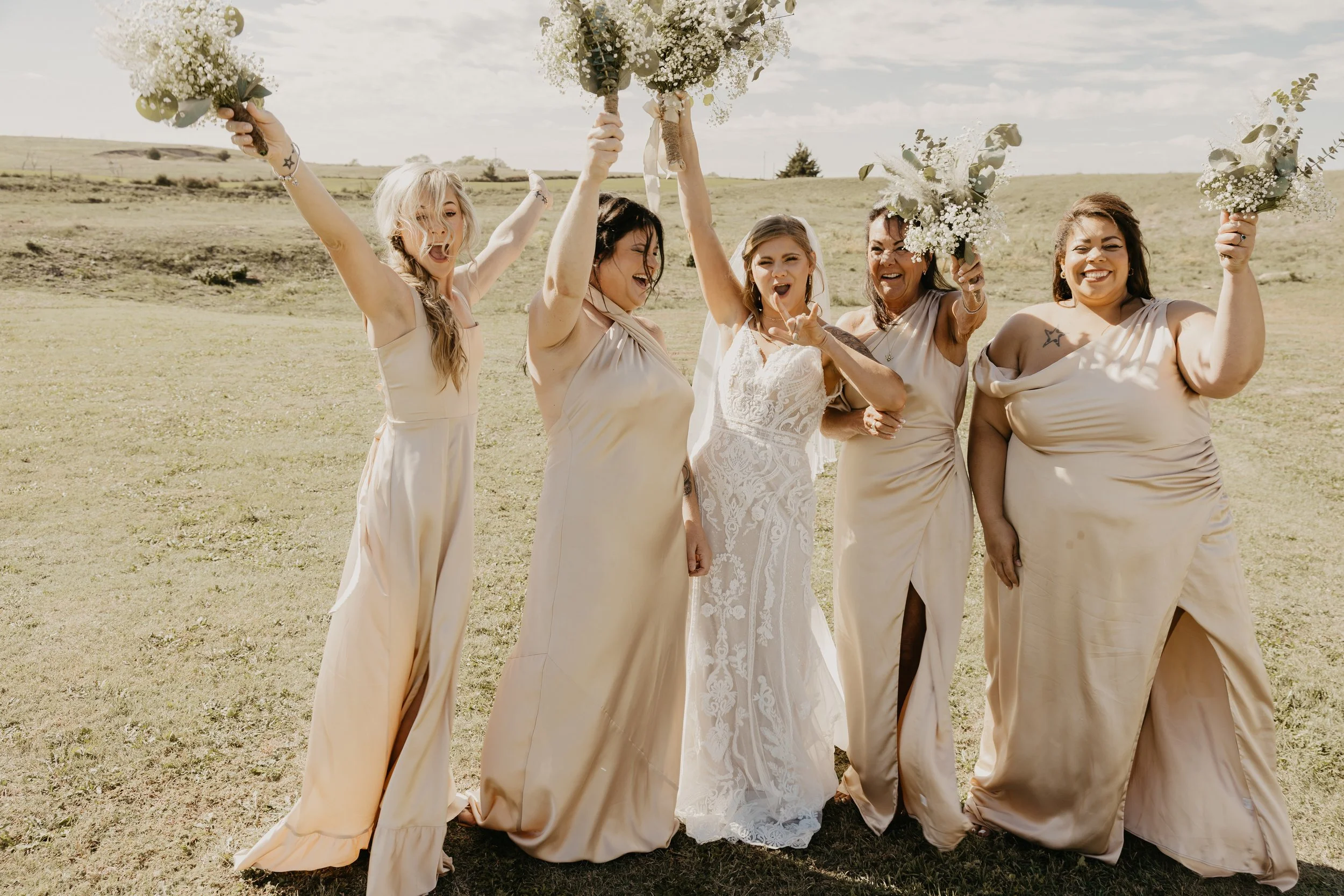 A bridal party celebrating outdoors at a wedding, holding bouquets and cheering on a sunny day.