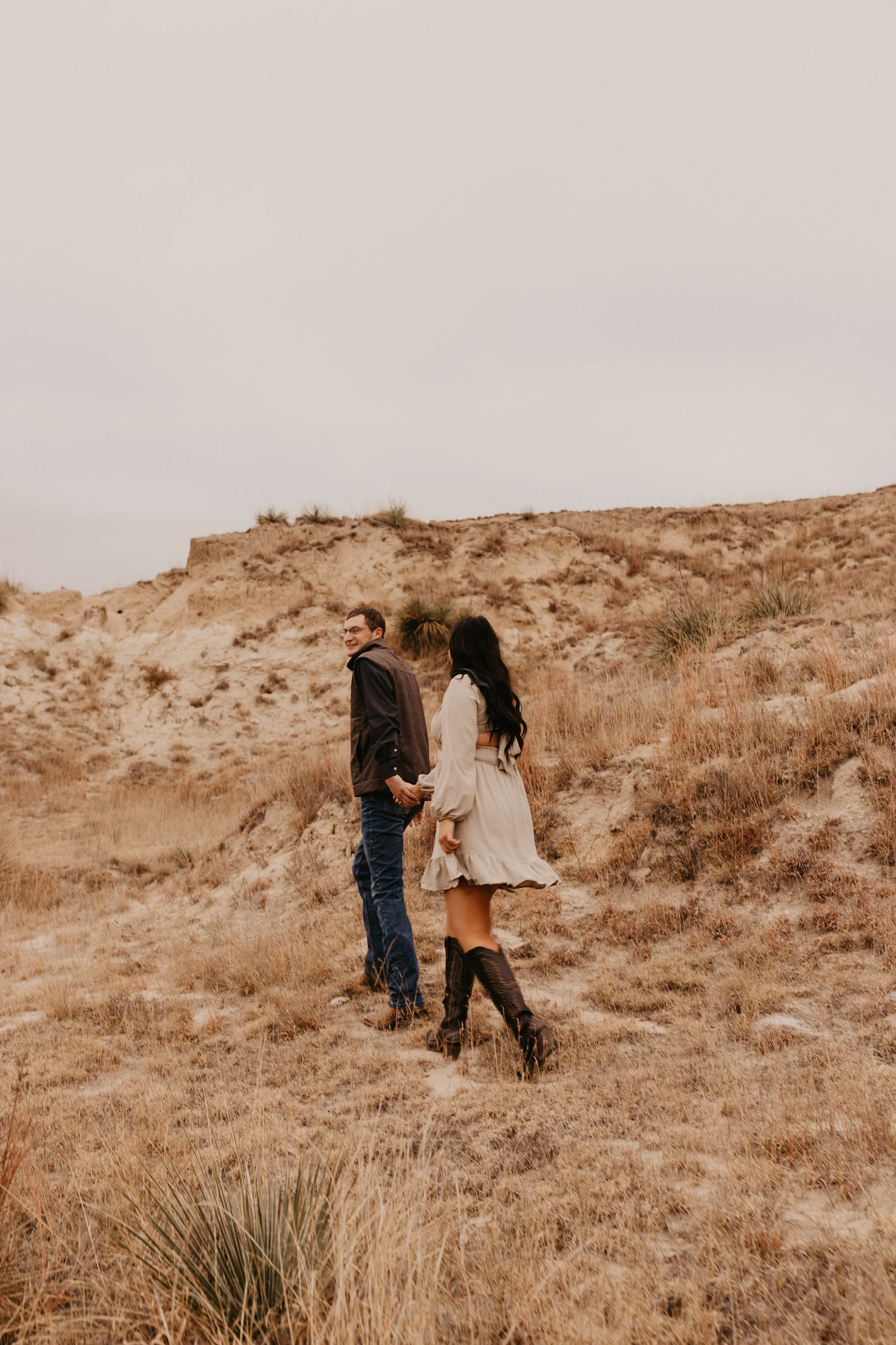 A man and woman holding hands while walking up a desert hillside with dry grass and sparse vegetation, overcast sky in the background.
