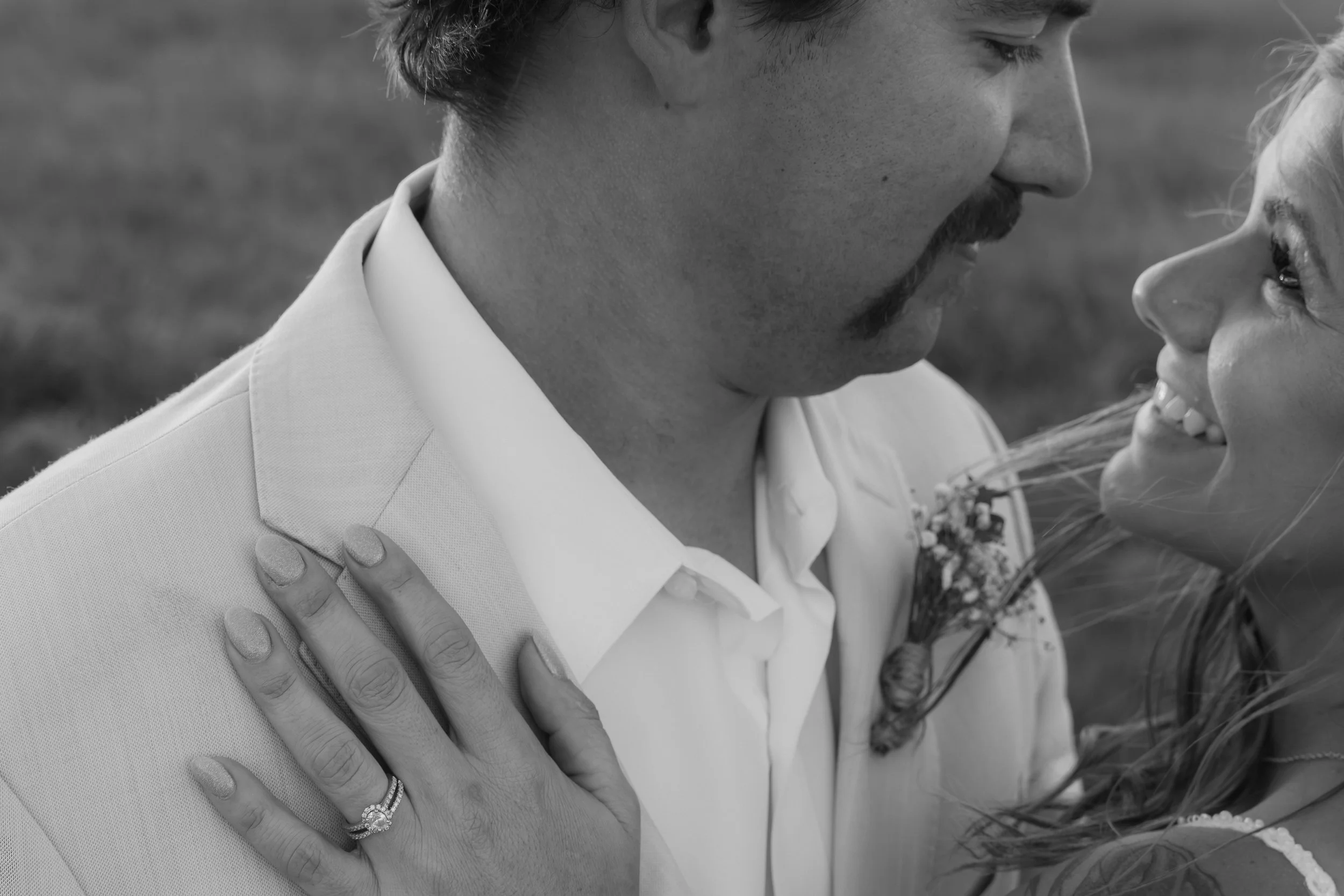 A black and white photo of a groom and bride close together, smiling at each other. The woman has a ring on her finger and her hand resting on the man's shoulder.