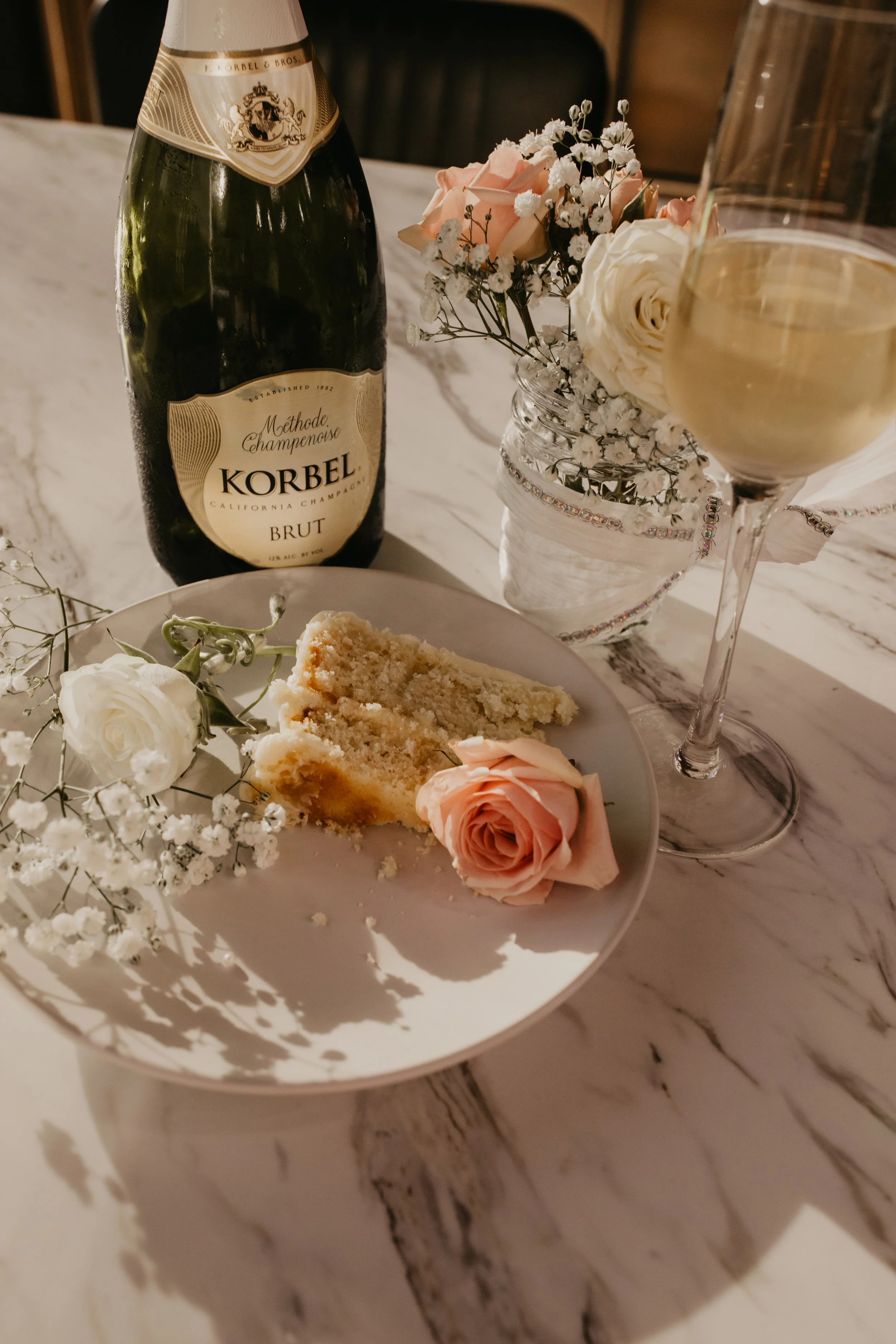 A slice of cake on a white plate with pink and white roses and baby's breath flowers, a glass of champagne, a bottle of Korbel champagne, and a vase with similar flowers on a marble table.