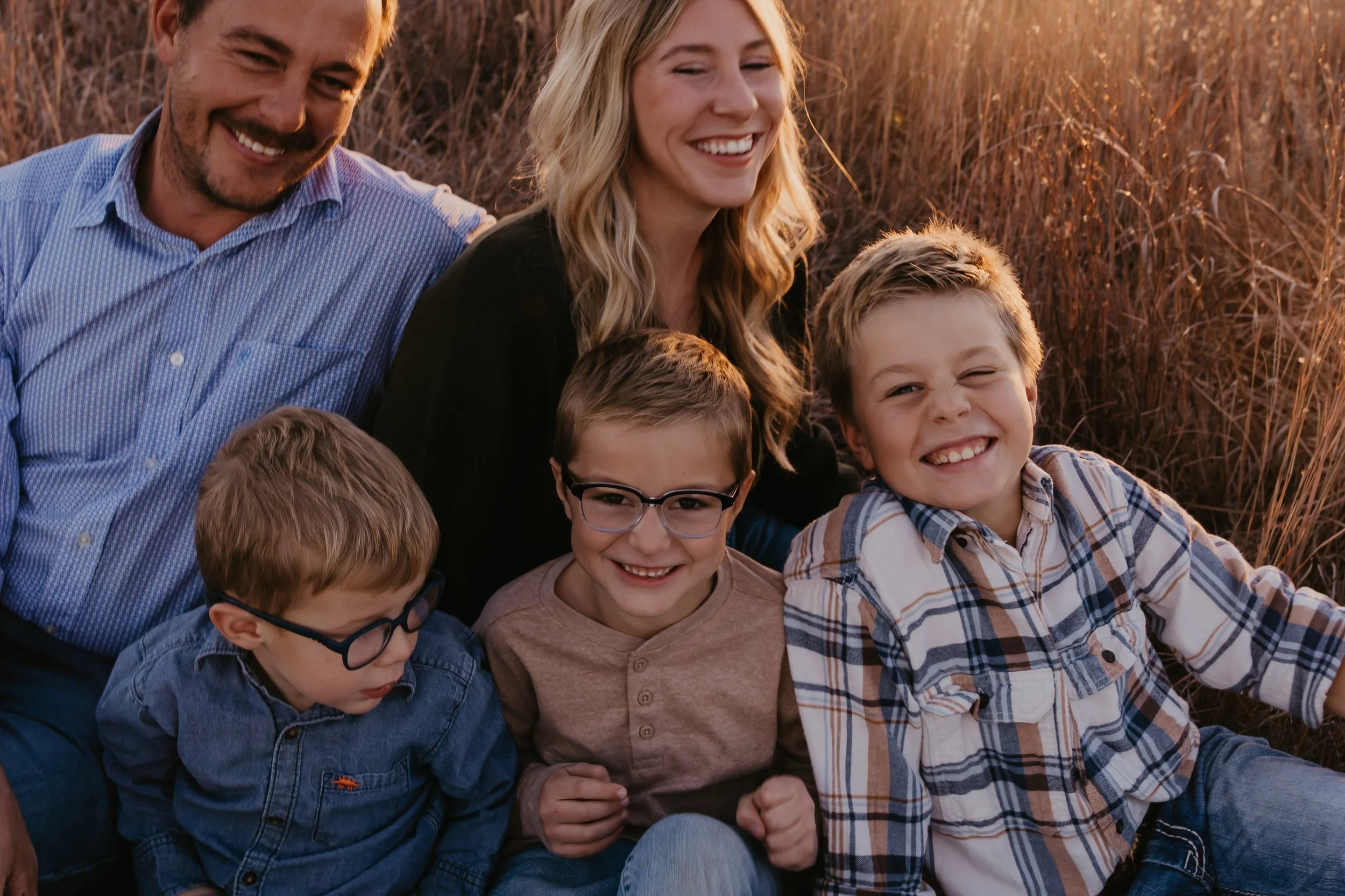 A family of five, including two adults and three children, sitting and smiling in a field during sunset.
