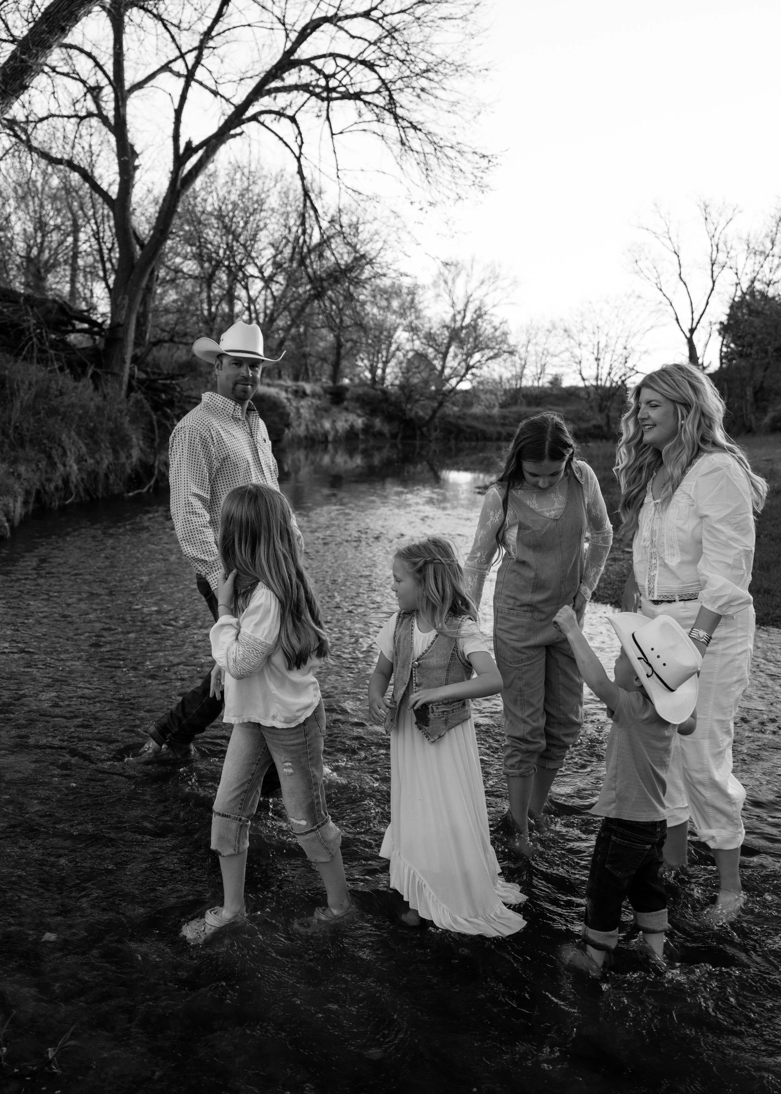 A family of six, including two adults and four children, standing in a shallow creek or river, enjoying a moment outdoors near leafless trees with a bright sky overhead.