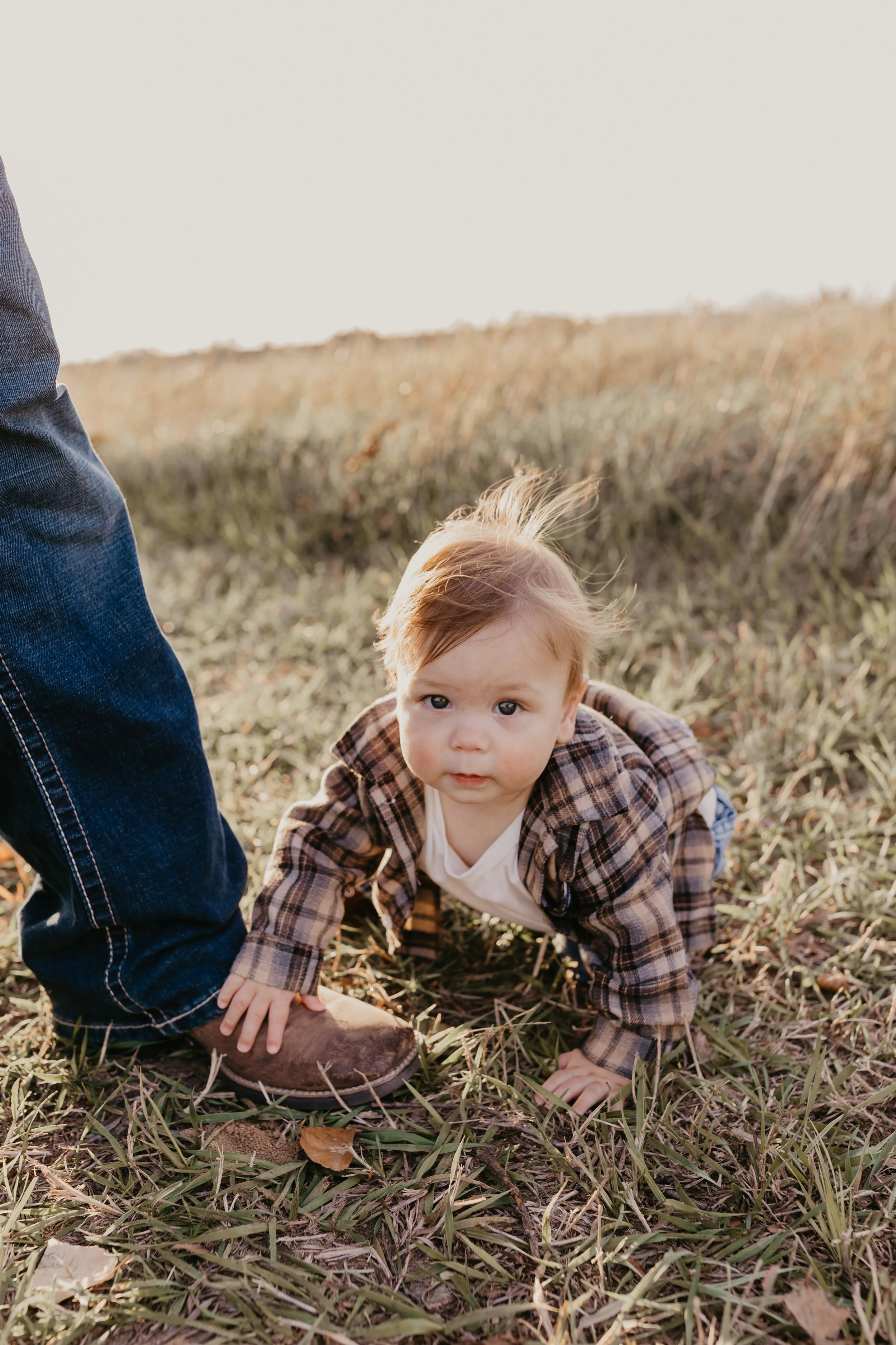 A young child crawling on the grass outdoors, with an adult partially visible standing nearby. The child is wearing a plaid jacket and has light brown hair.