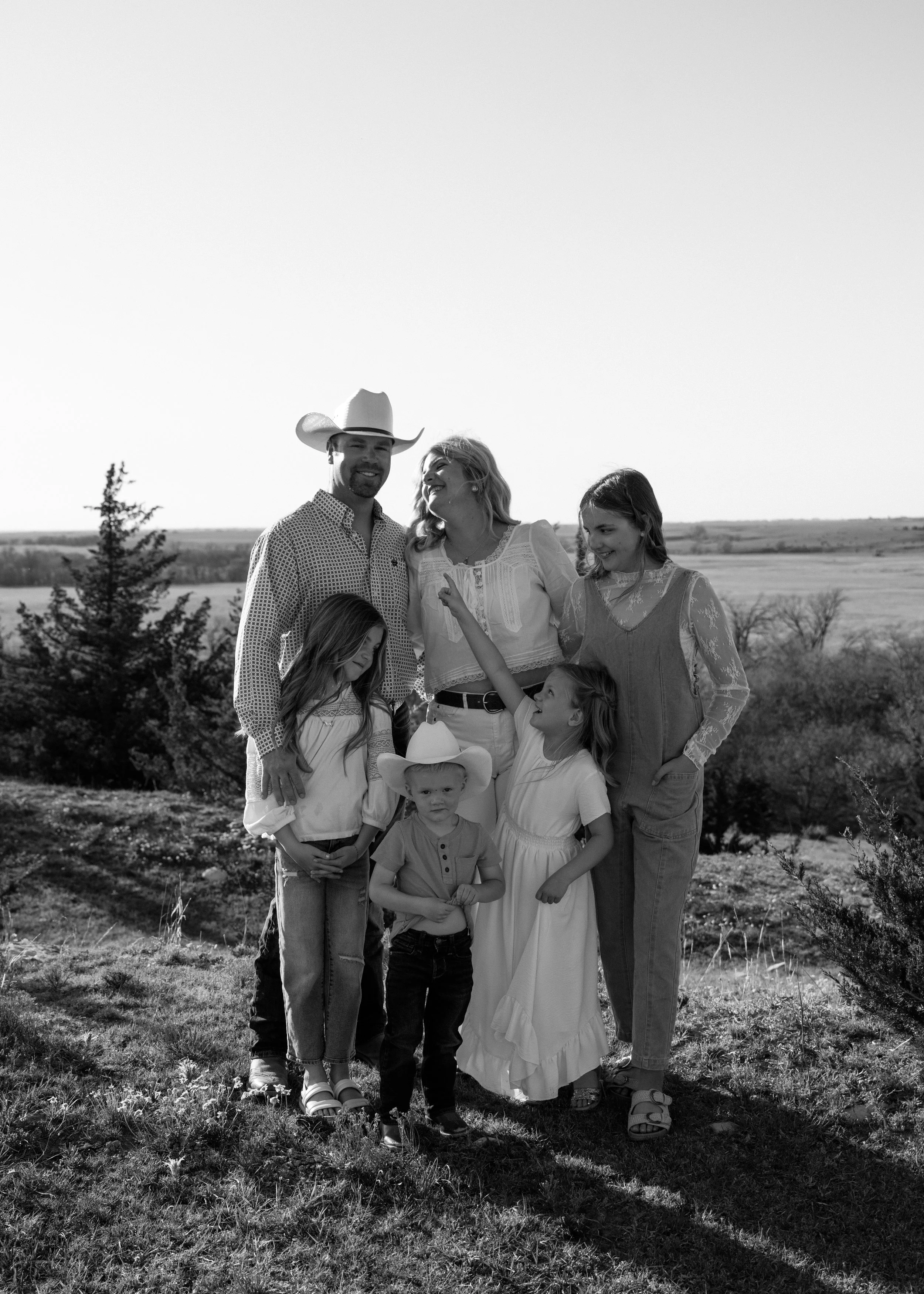 A family of six standing outdoors in a rural setting, wearing casual and western-style clothing, with some children smiling and interacting with each other.
