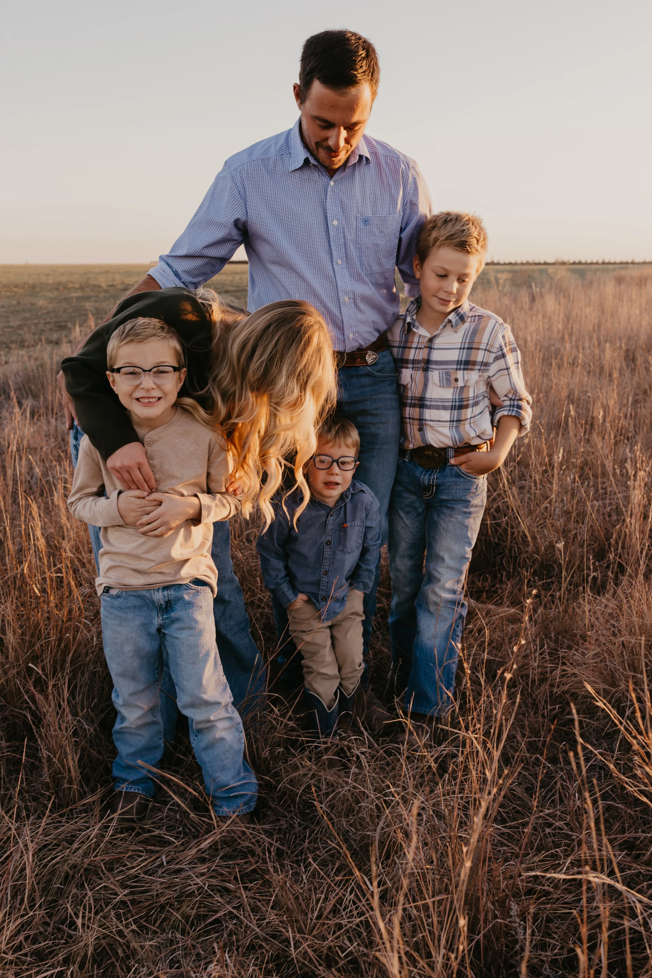 Parents and their three children standing in a field of tall grass during sunset, all smiling and looking down.