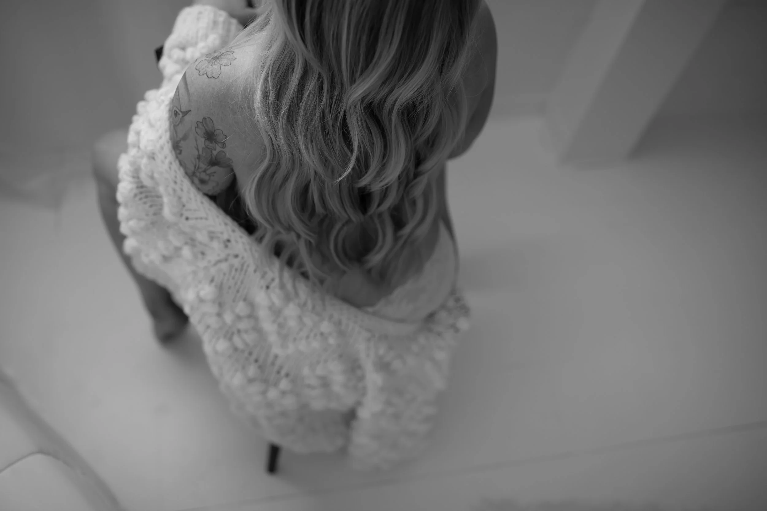 Top-down black-and-white photo of a woman with long wavy hair, wearing a knitted sweater with lace details on shoulders, seated on a chair in a minimalist room.