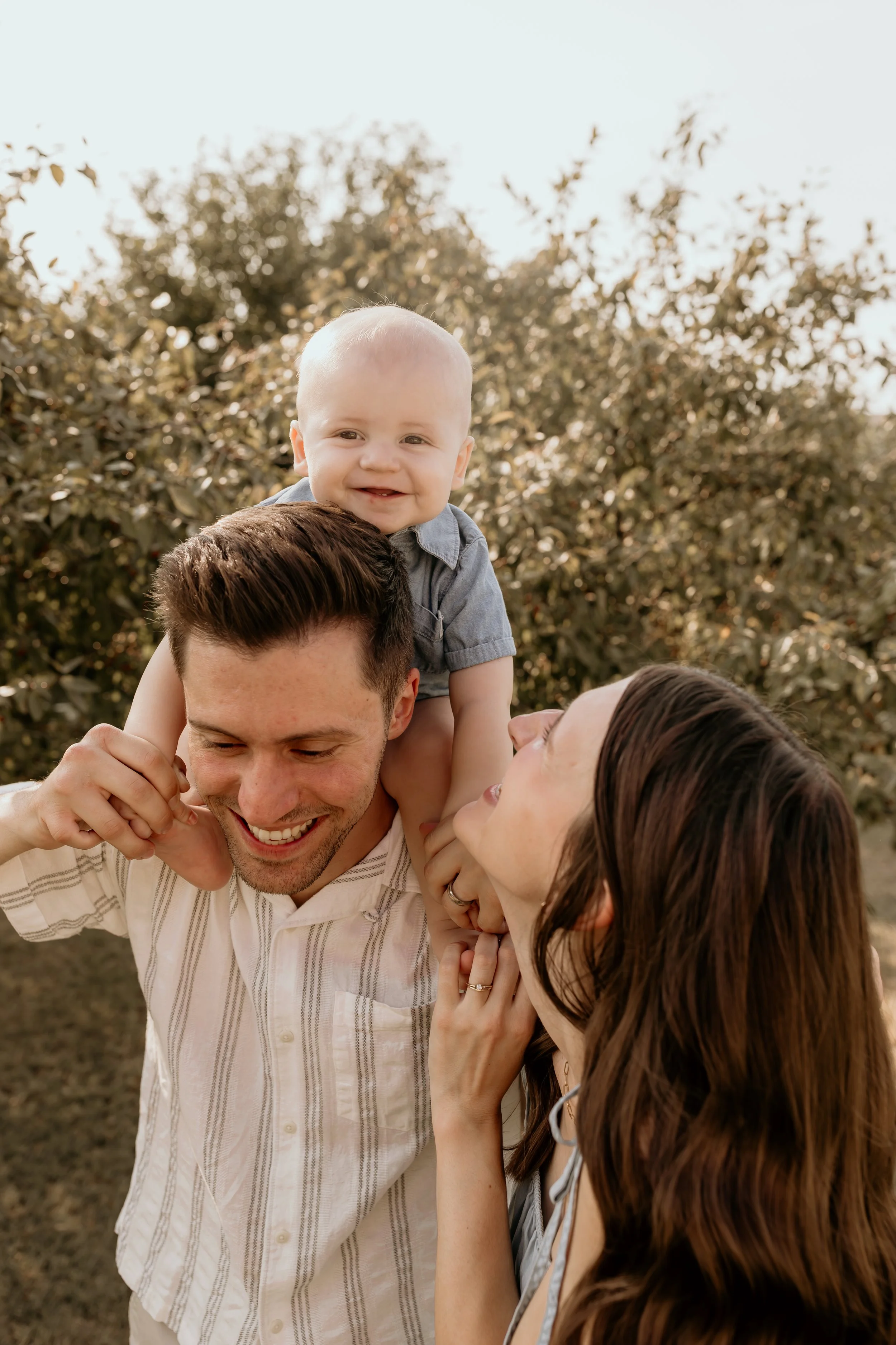 A happy family outdoors, with a man carrying a smiling baby on his shoulders, while a woman looks up at them affectionately.