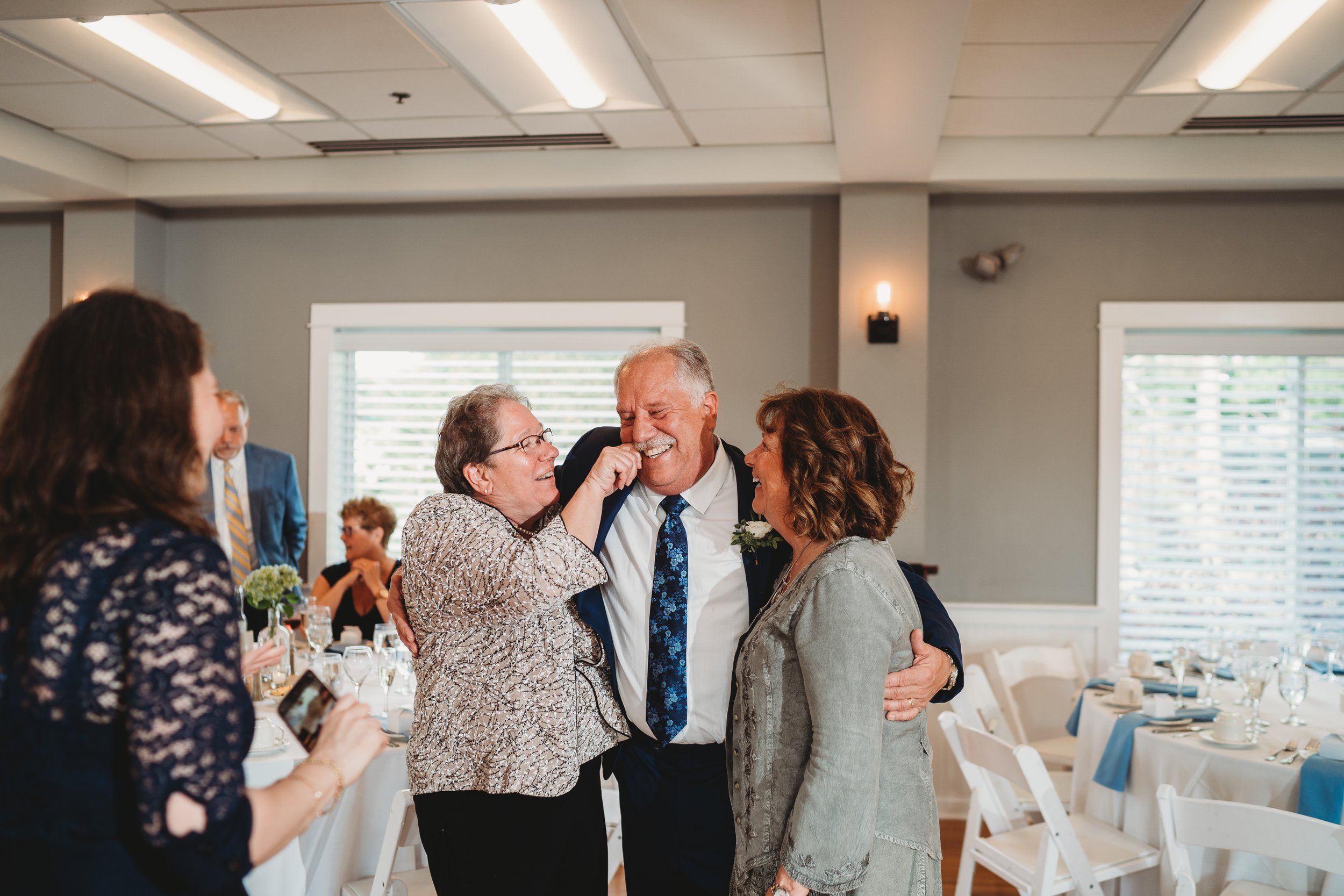 Group of people at a celebration, with three individuals hugging and laughing in the center, inside a decorated event hall with tables set for a meal.