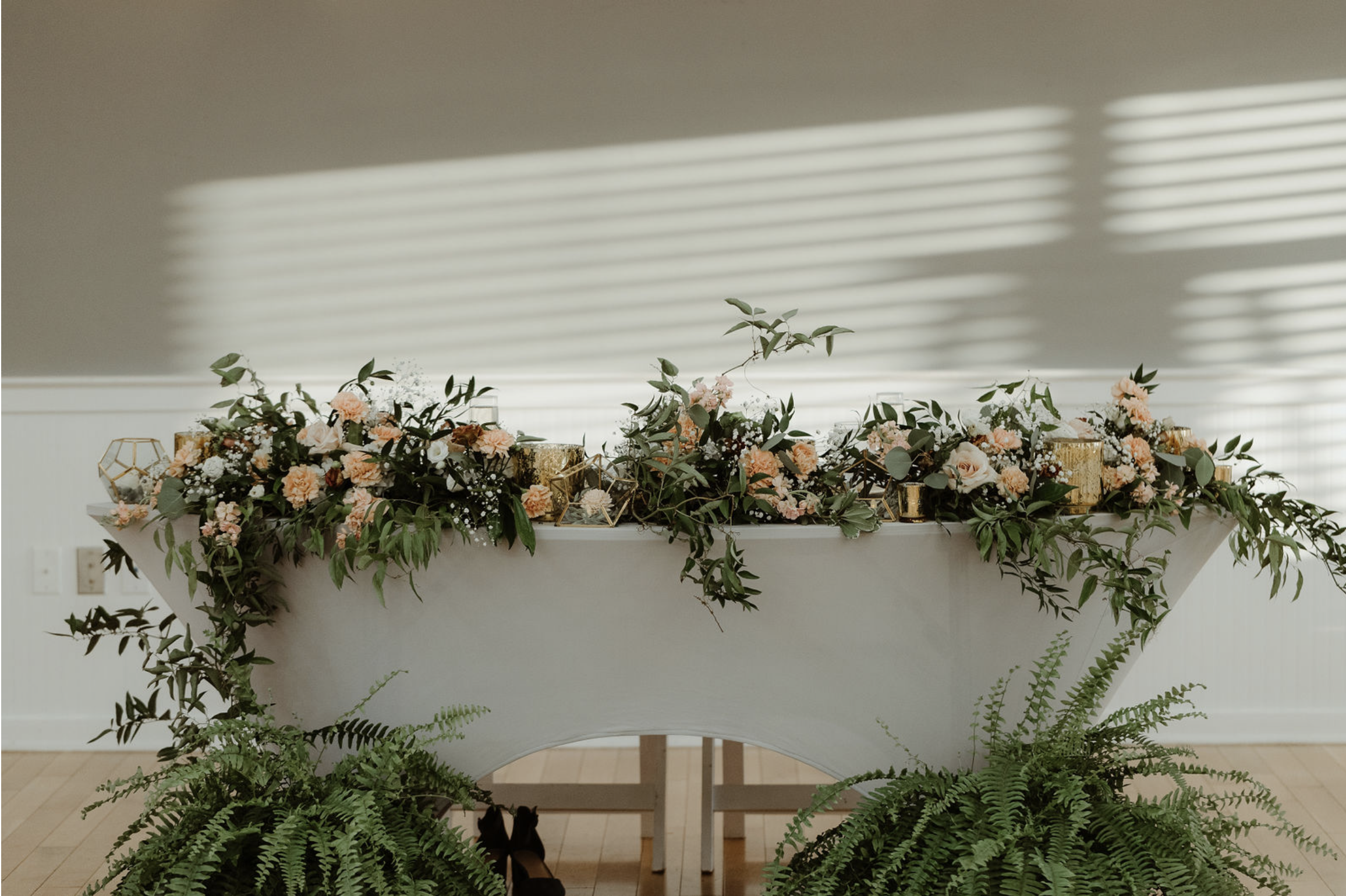 Elegant white table decorated with pink and white flowers, green foliage, and gold geometric accents, set against a neutral background with striped sunlight patterns.