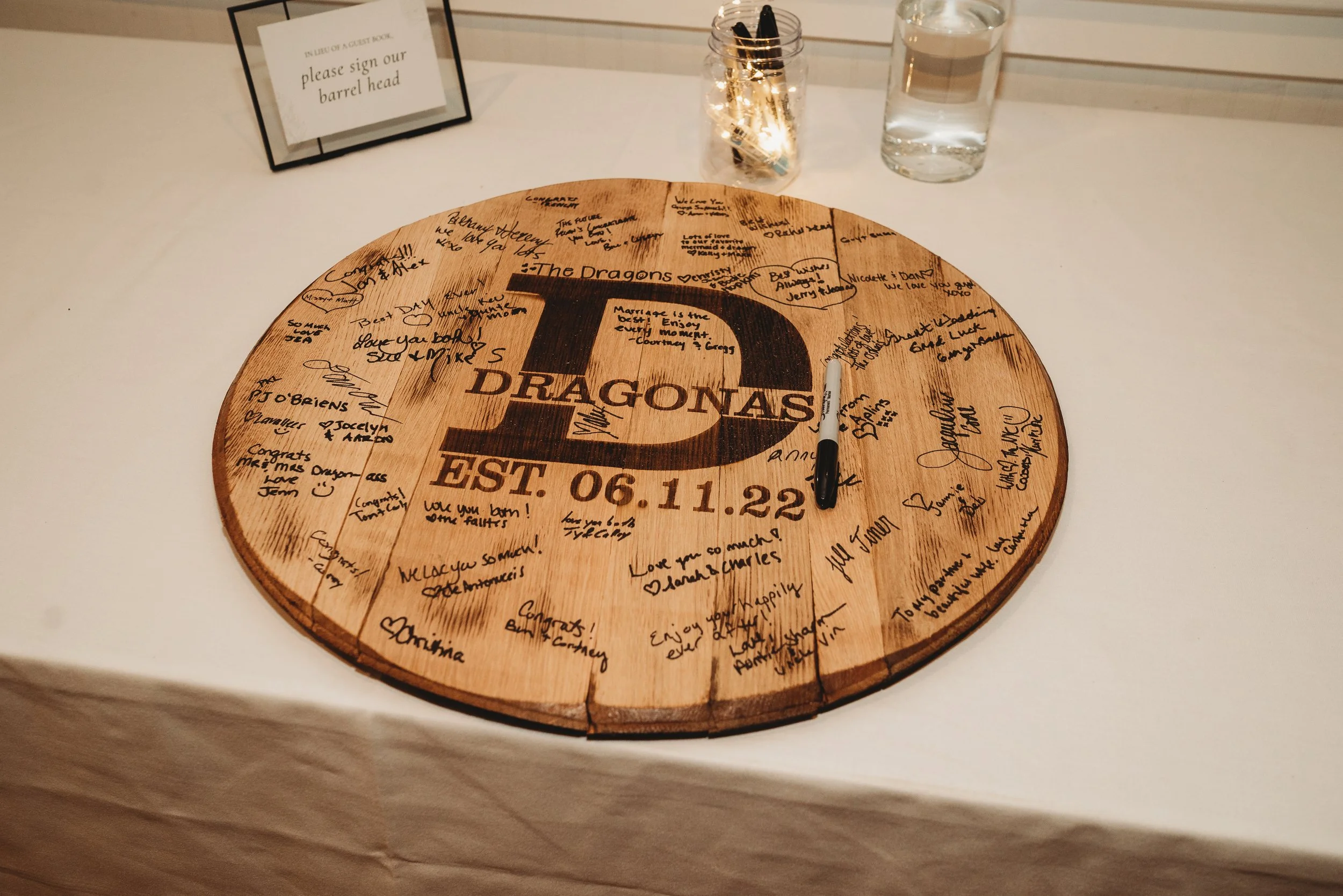 Round wooden table with the name 'The Dragons' and the date 'EST. 06.11.22' engraved on the surface, covered with handwritten messages and signatures. There is a black marker resting on the table, a glass container with string lights and pens behind 