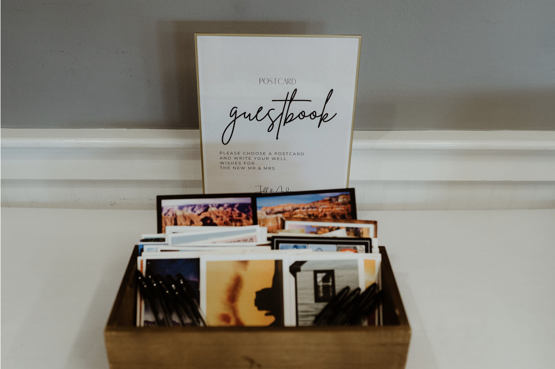 A wooden box filled with postcards placed on a white table in front of a white wall. Behind the box, there is a white sign with gold border that reads "POSTCARD guestbook" and instructions to write well wishes for the new Mr. and Mrs.