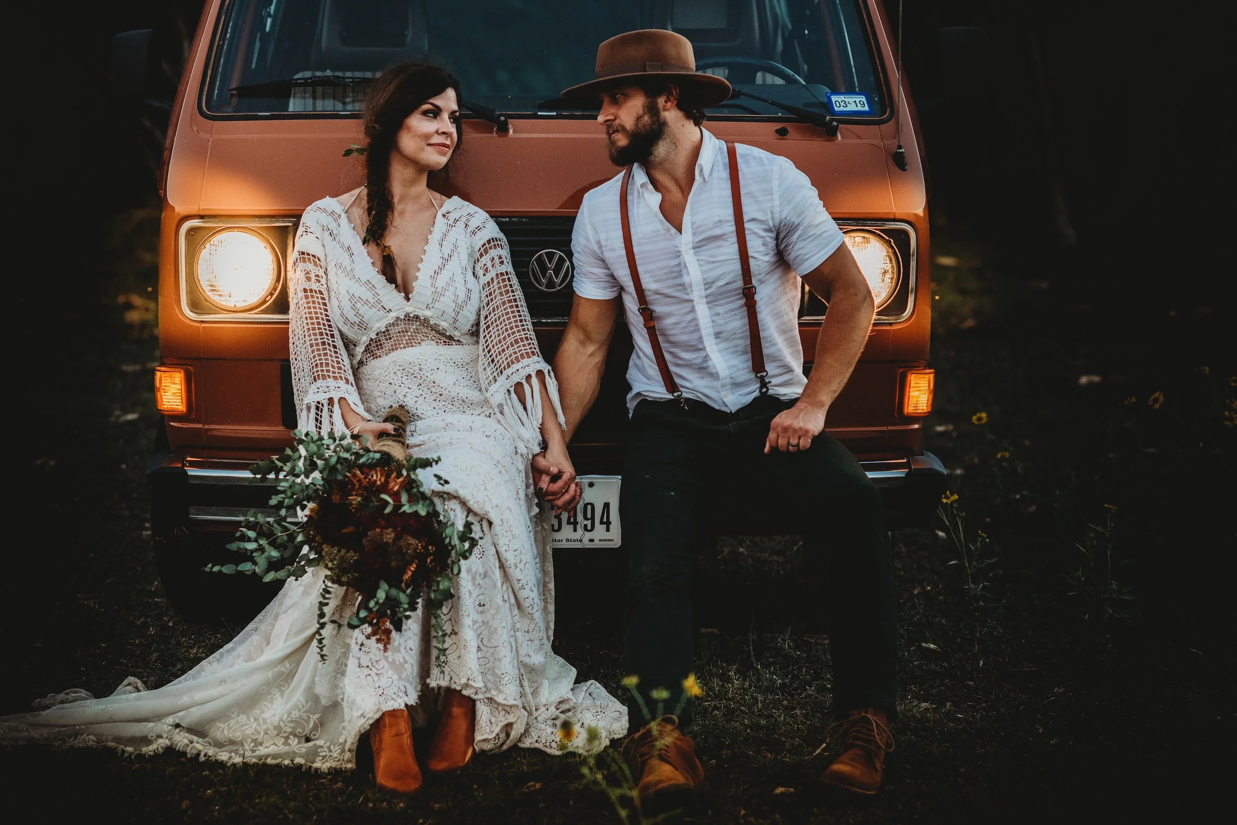 A woman in a boho white lace wedding dress with brown boots, holding a bouquet, sitting on the front of an orange Volkswagen van, holding hands with a man in a white shirt, suspenders, and a brown wide-brimmed hat, sitting on the bumper. They are outdoors during sunset, surrounded by small yellow flowers, with a dark background.